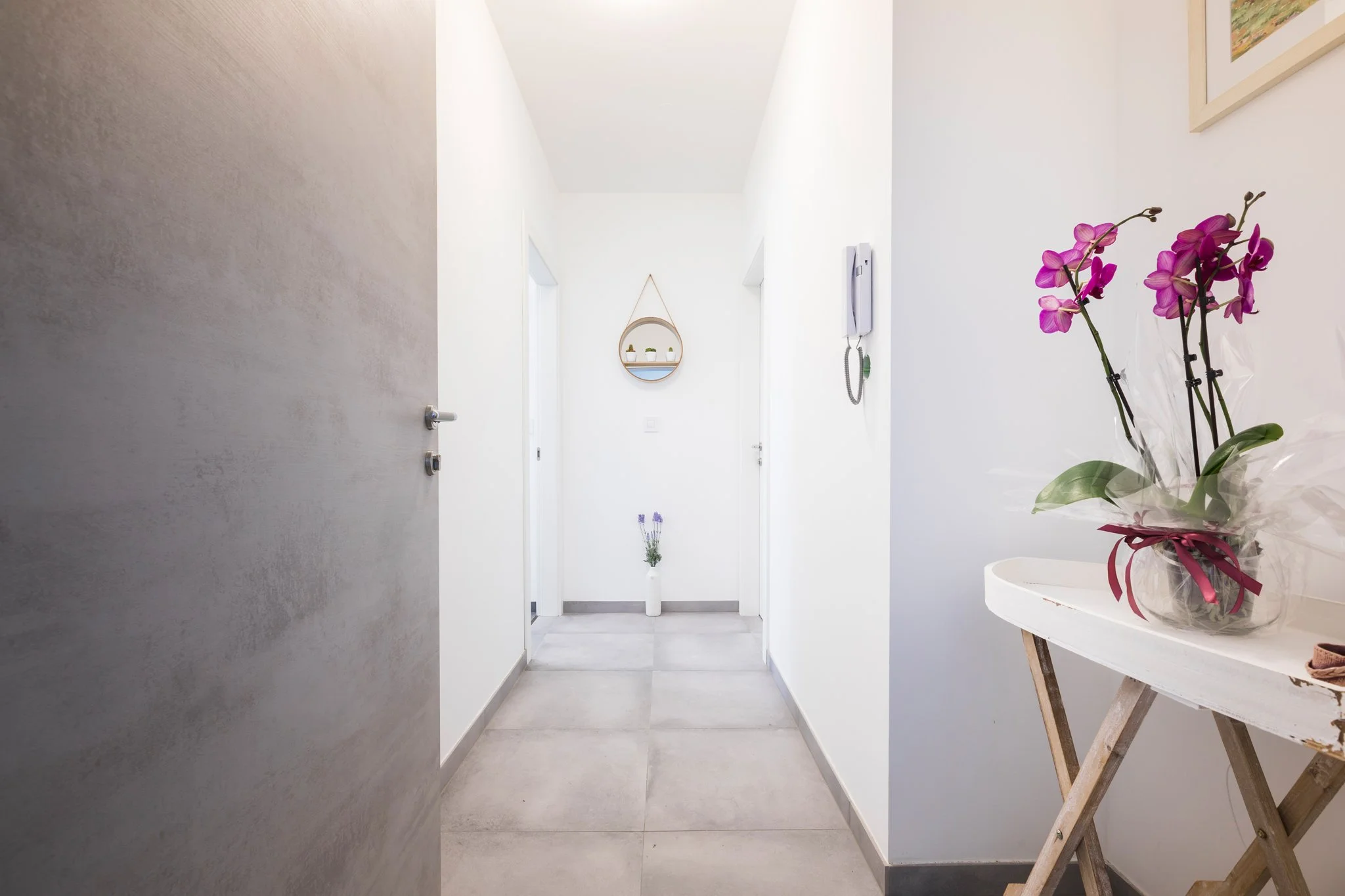 Interior view of a minimalistic hallway with white walls and gray tiled floor, decorated with a wall-mounted round shelf with small potted plants and a hallway flower arrangement of purple orchids on a small table.