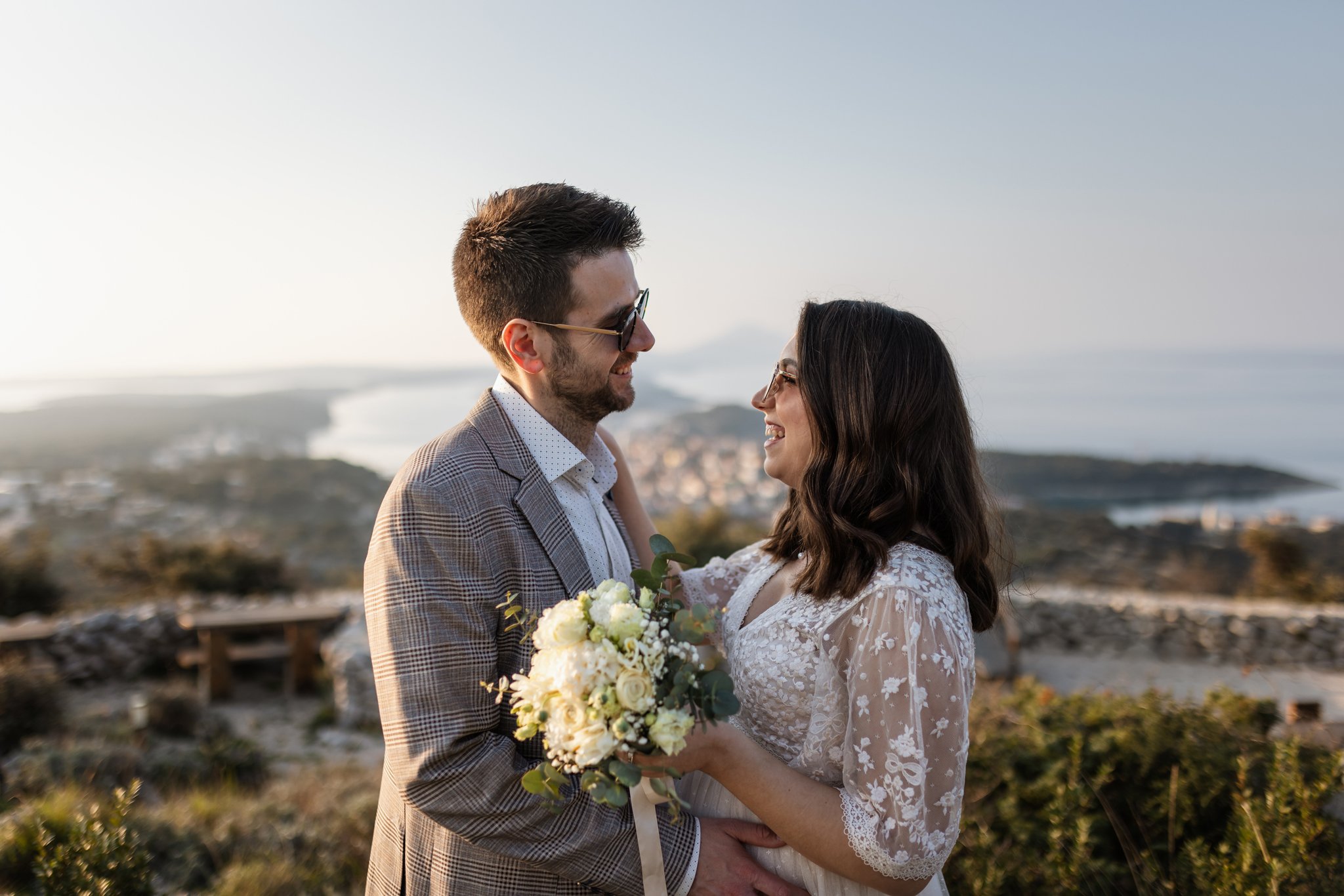A couple dressed in wedding attire smiling and looking into each other's eyes outdoors, with the woman holding a bouquet of white flowers.