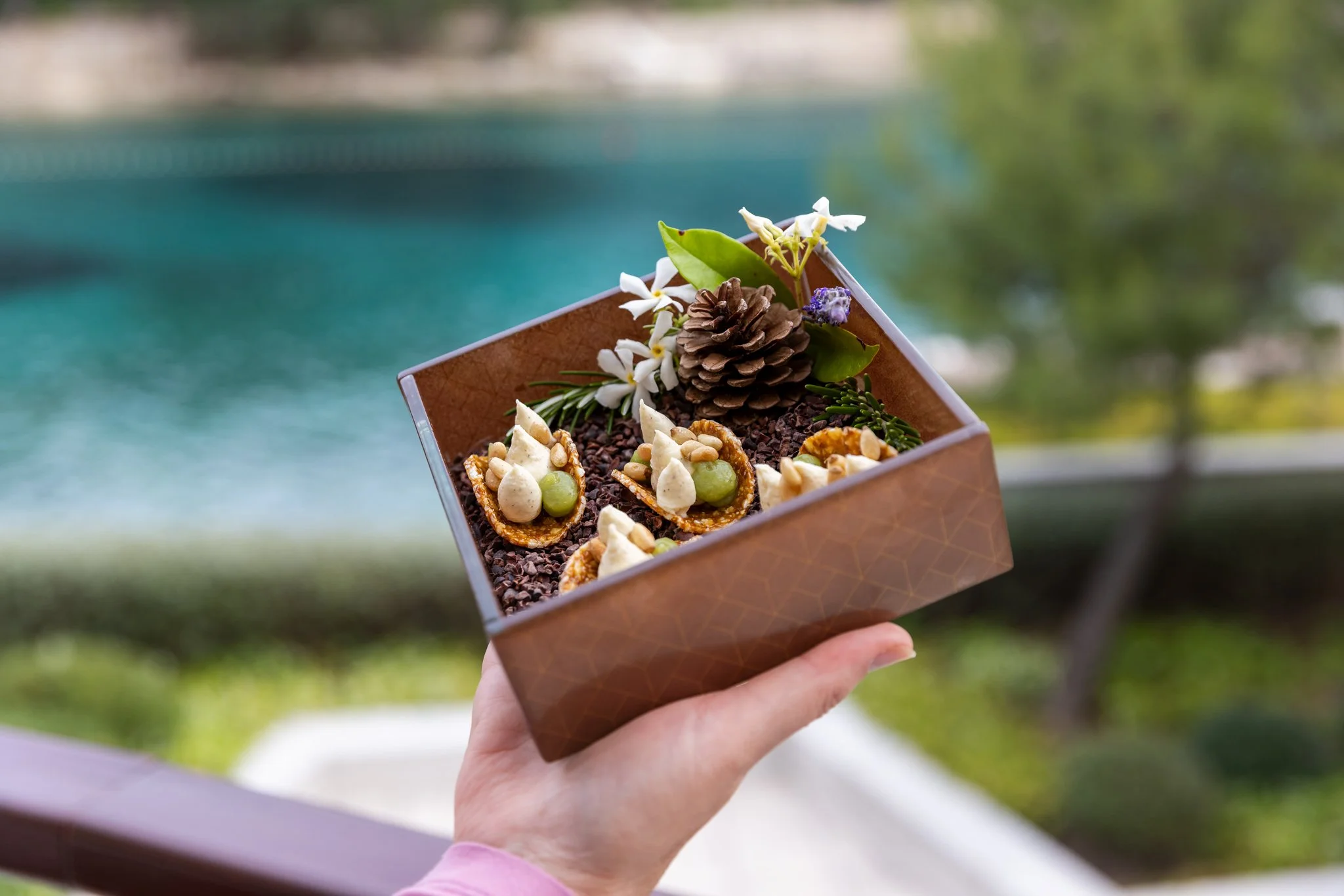 A hand holding a square container filled with small decorative items, flowers, pinecone, and greenery, with a blurred outdoor background.