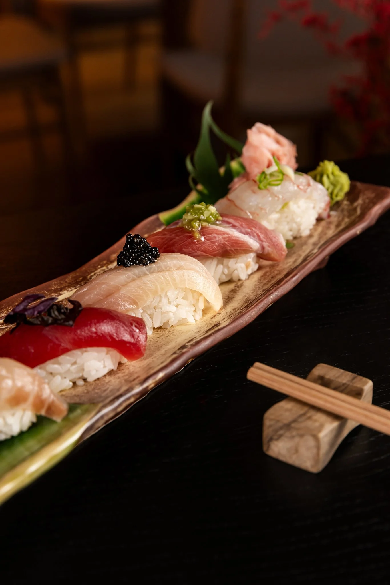 A long rectangular wooden plate with various pieces of sushi, topped with different garnishes, against a dark background with small wooden chopsticks.