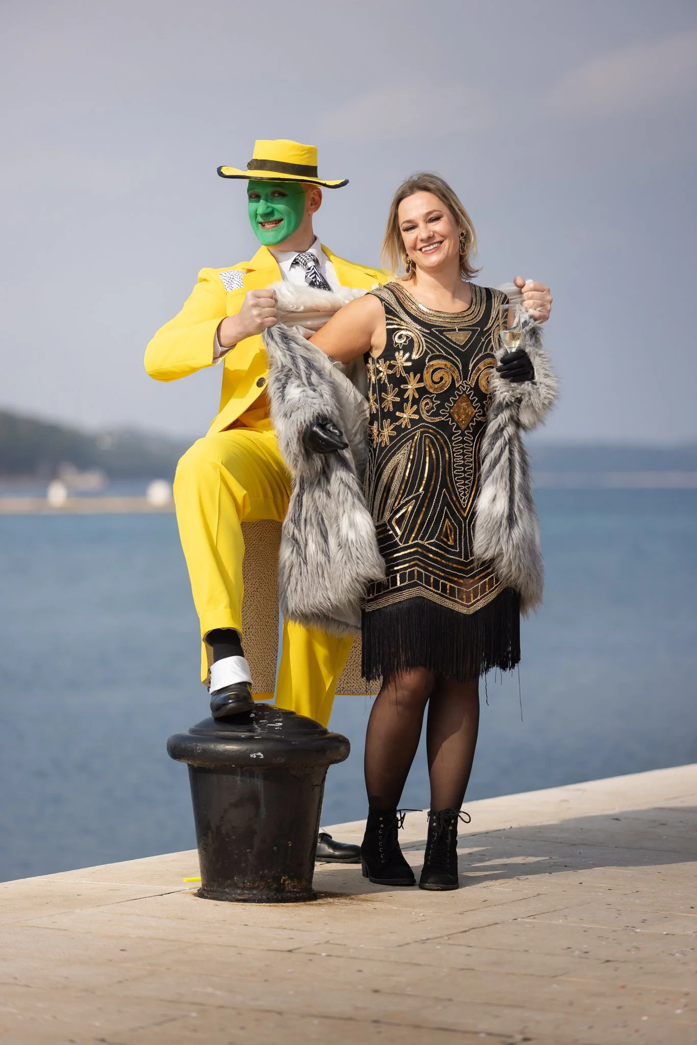 Man dressed as a green-faced character in a bright yellow suit and hat, standing behind a woman in a black and gold dress with a fur coat, holding a glass of champagne, by a waterfront.