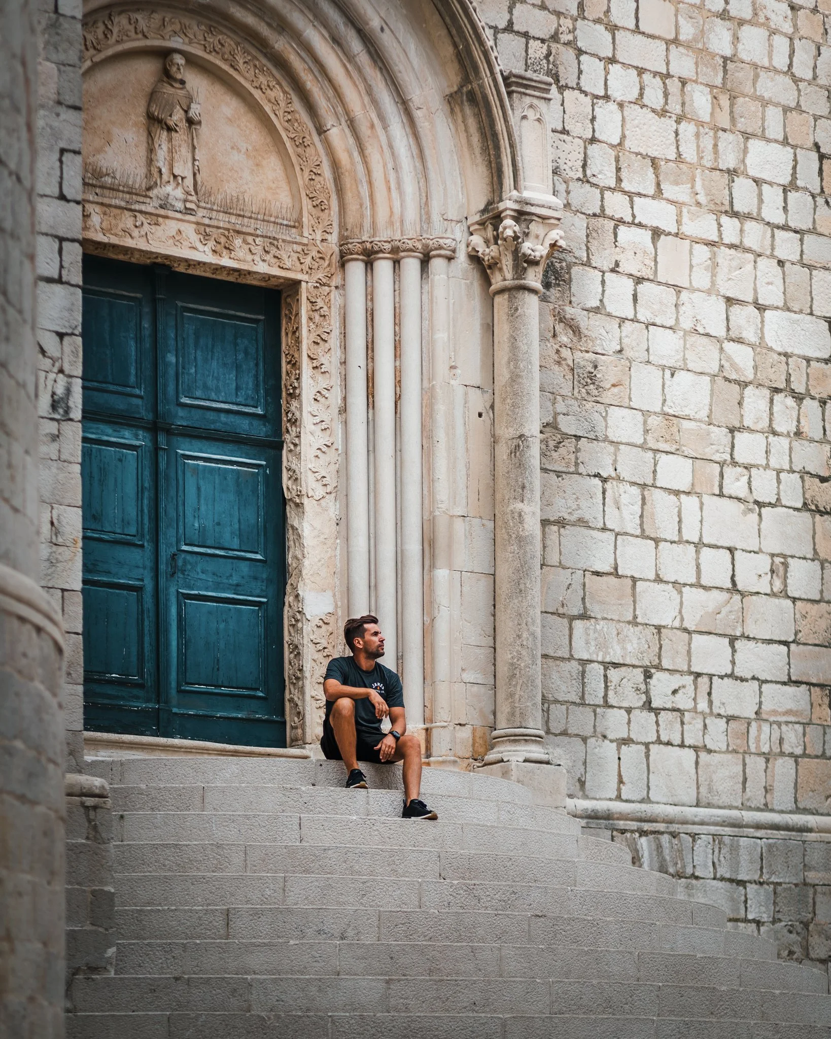 A man sitting on the steps of a historic stone building with ornate architectural details, including carved columns and a carved relief of a priest or monk holding a cross, in a relaxed pose.