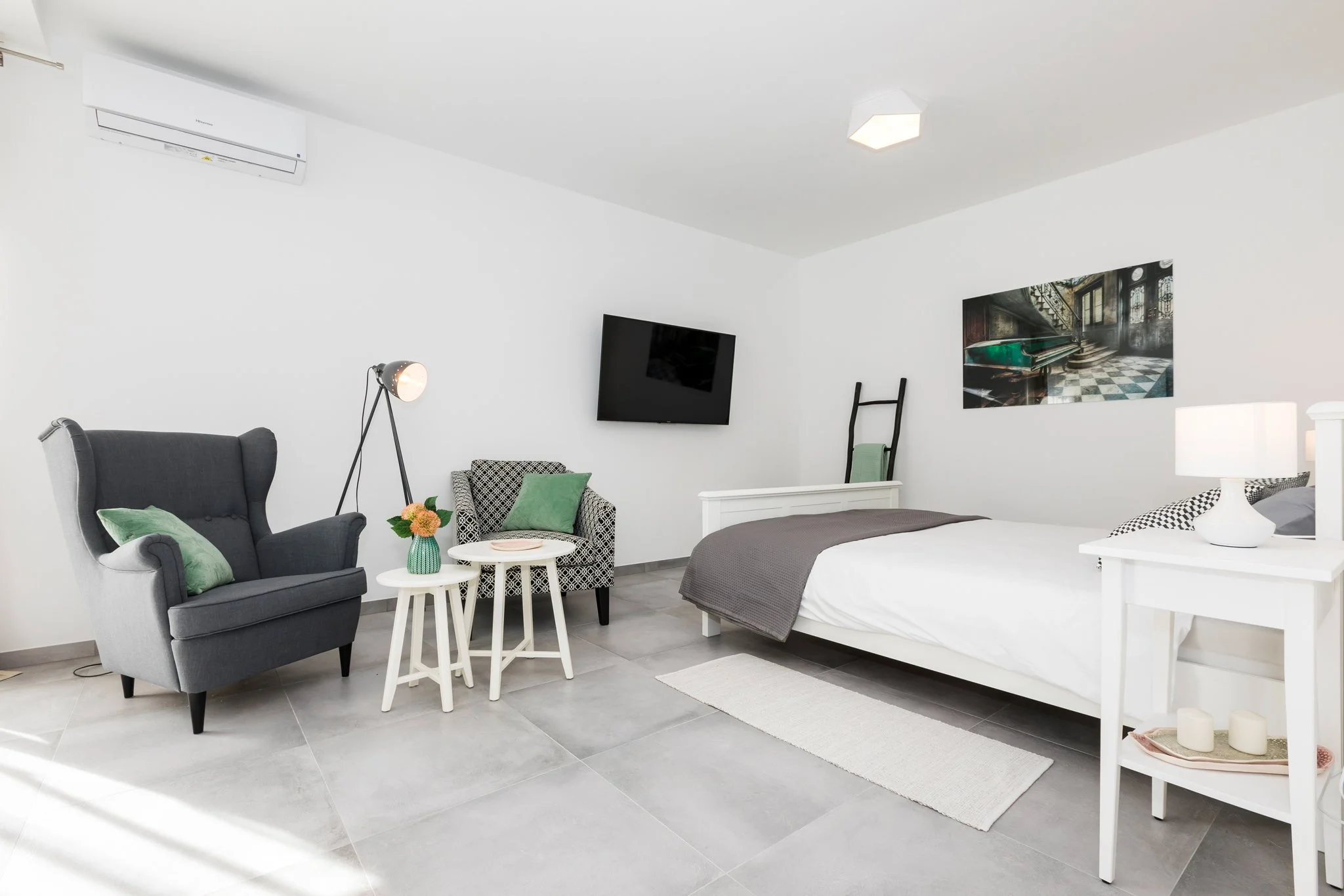 Modern, minimalistic bedroom with white walls and gray tile flooring, featuring a black armchair with green cushions, a small round white table with a vase of flowers, a wall-mounted flat-screen TV, a bed with white sheets and a gray blanket, and abs