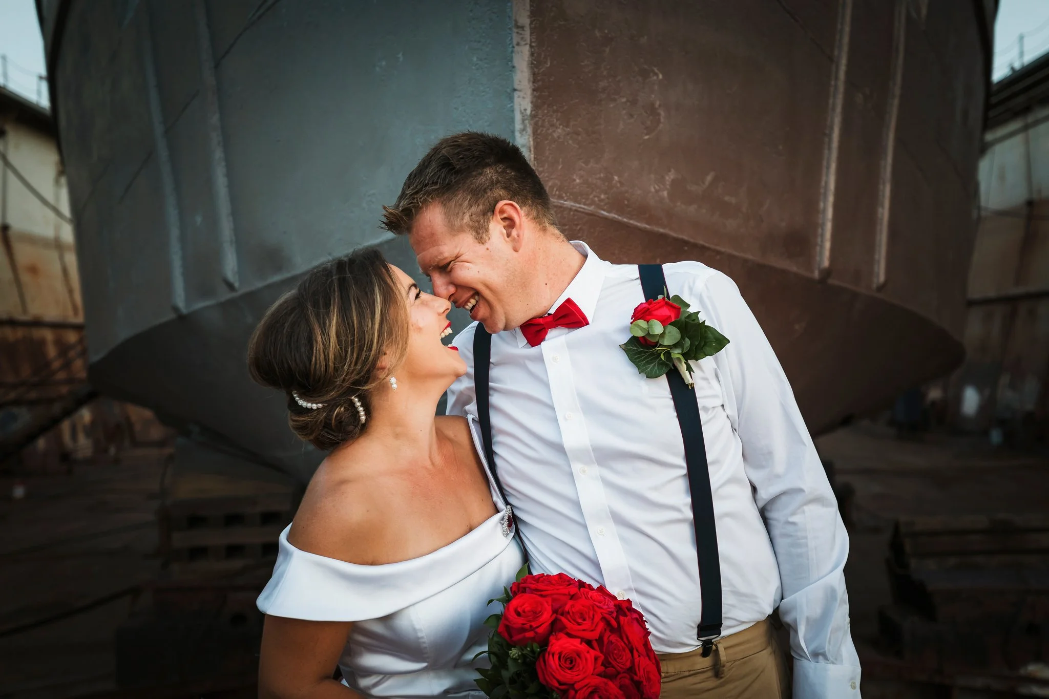 A joyful couple in wedding attire sharing a close moment outdoors, in front of a boat, with the woman holding a bouquet of red roses.