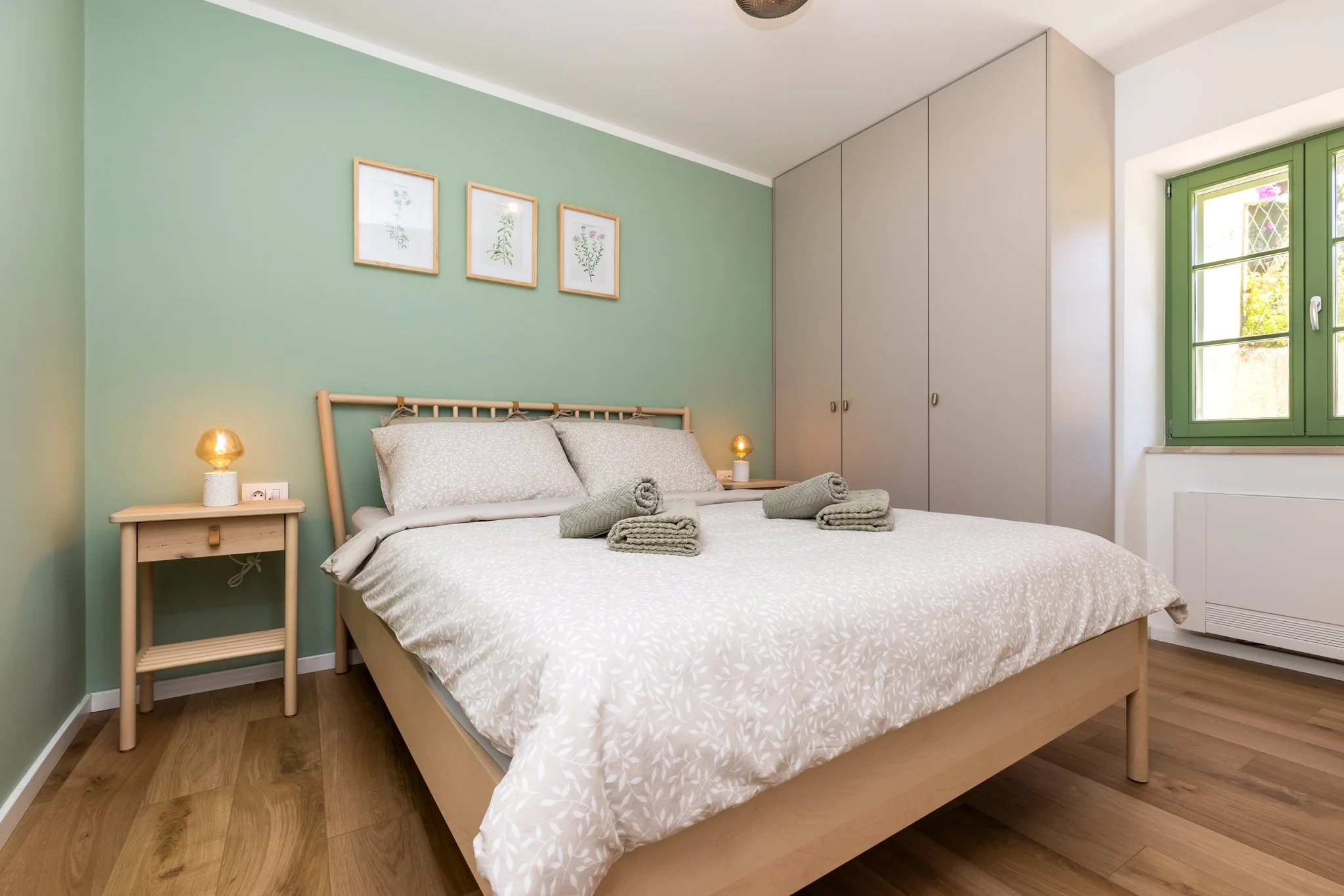 A cozy bedroom with a wooden bed, beige bedding, and rolled towels on the bed. There is a nightstand with a lamp on each side and framed botanical artwork above the bed. A large wardrobe is built into the wall, and a window with green framing shows s