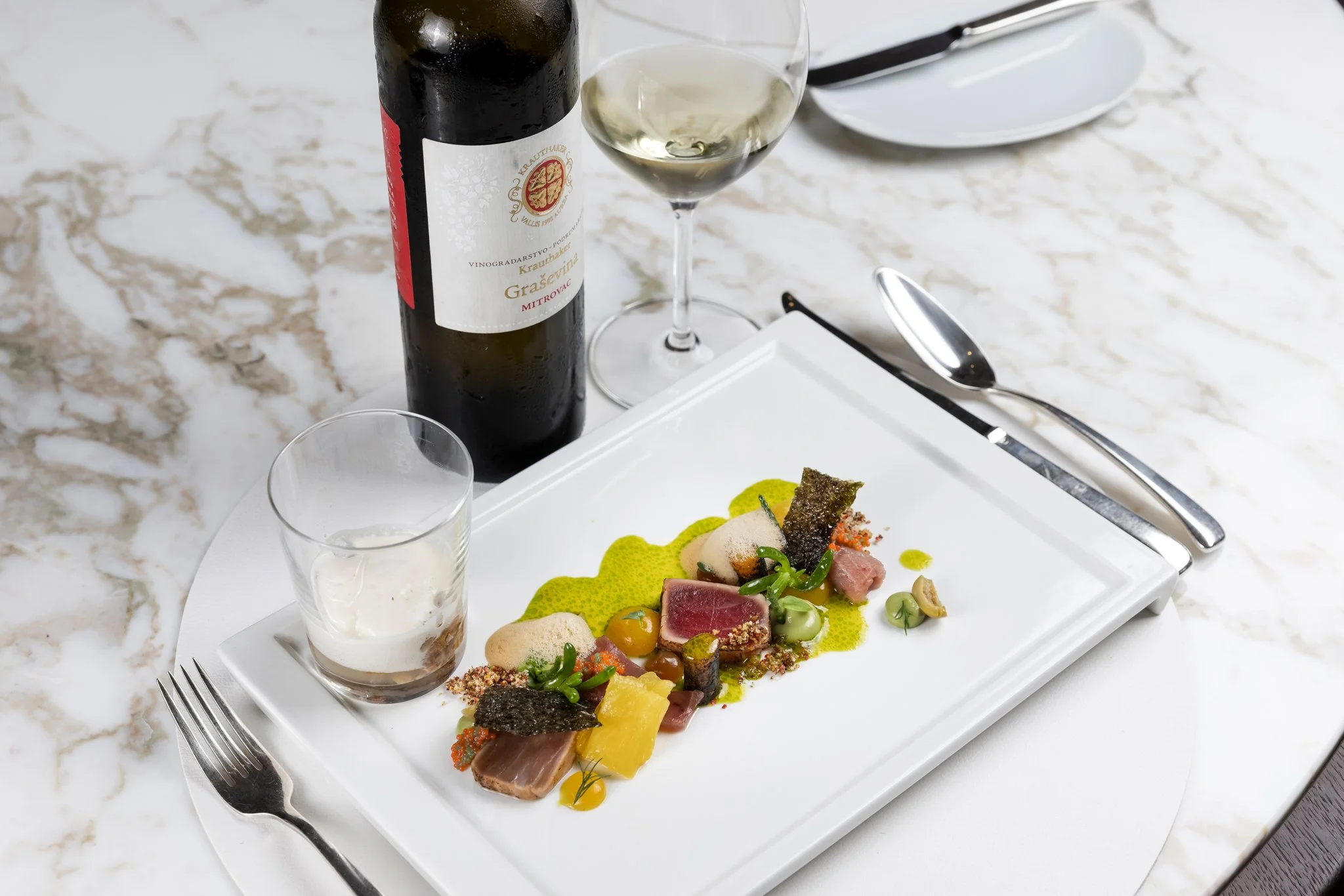 A white rectangular plate with assorted gourmet food items, drizzled sauces, and garnishes, with silver cutlery on a white marble table, and a bottle of white wine and a glass nearby.