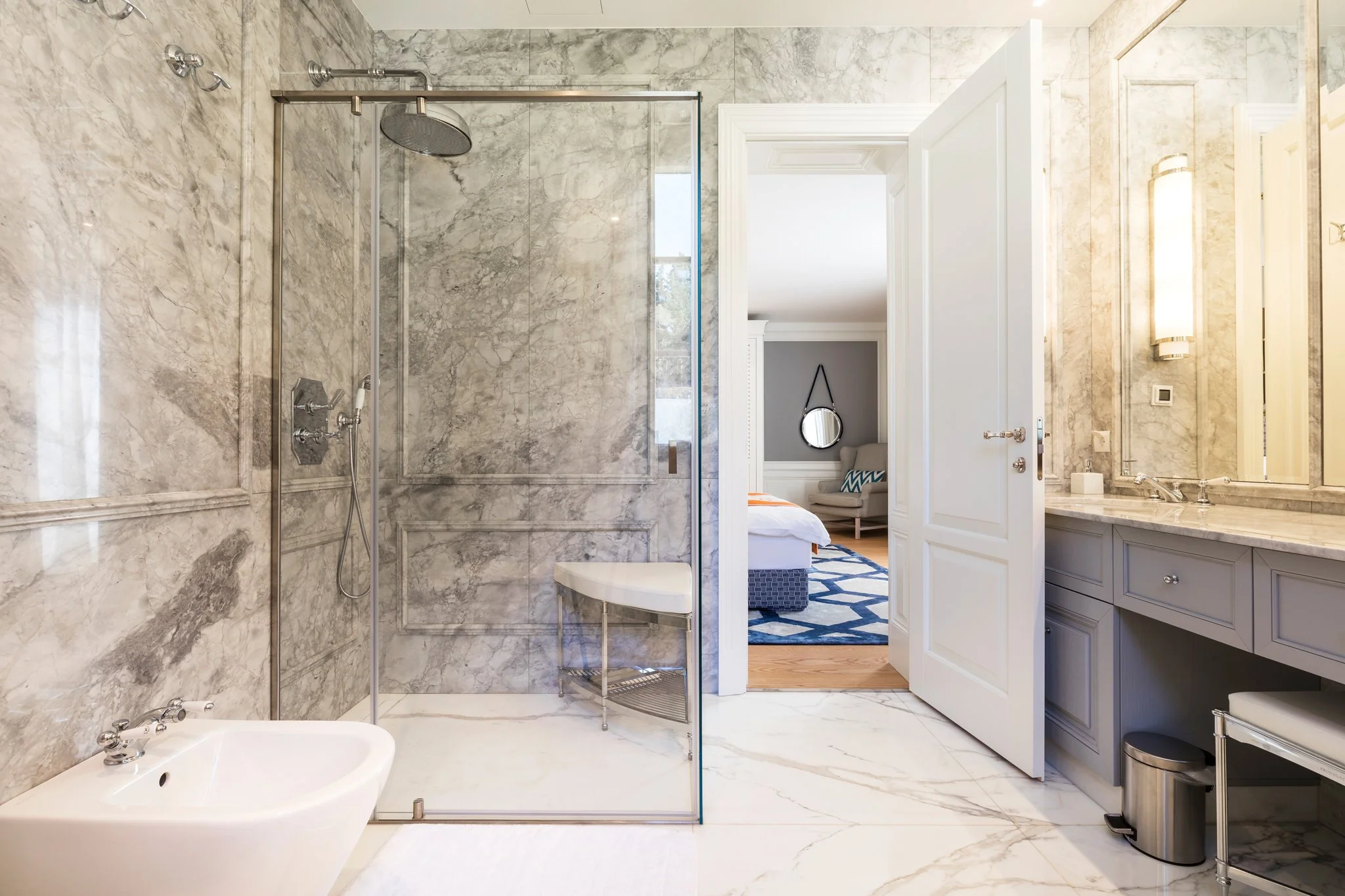 Bathroom with marble walls and floor, walk-in shower with glass door, toilet, sink with marble countertop, a mirror, wall-mounted light fixture, and a view into an adjacent bedroom with a chair and a small round mirror.