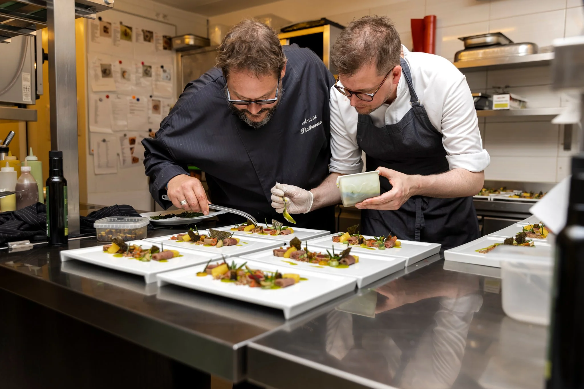 Two chefs in a commercial kitchen numbering and plating dishes with various garnishes and ingredients on white rectangular plates.