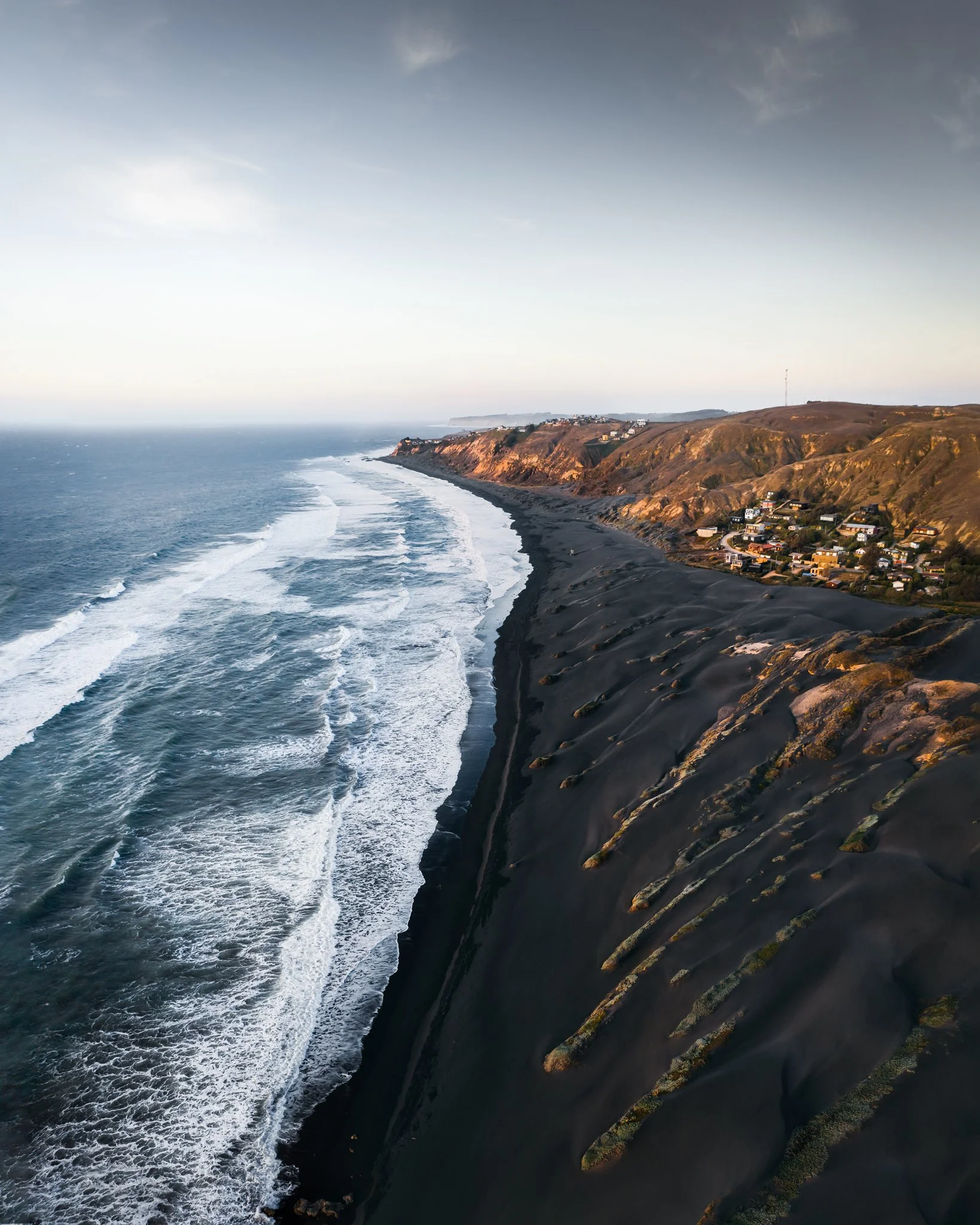 Aerial view of a rugged coastline with dark volcanic sand beaches, cliffs, and ocean waves washing ashore, with residential houses on the hillside.