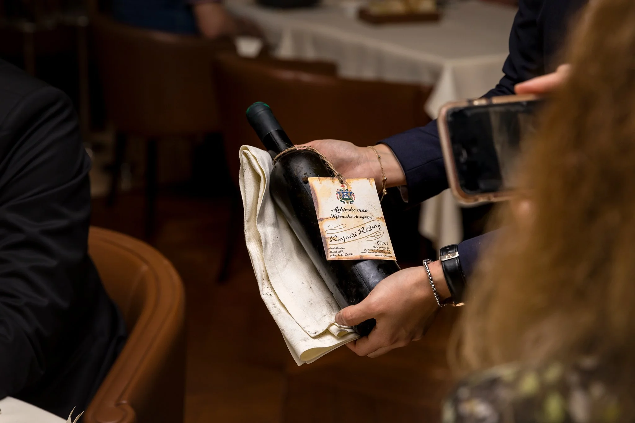 Person holding a wine bottle with a label in a restaurant setting, surrounded by other people