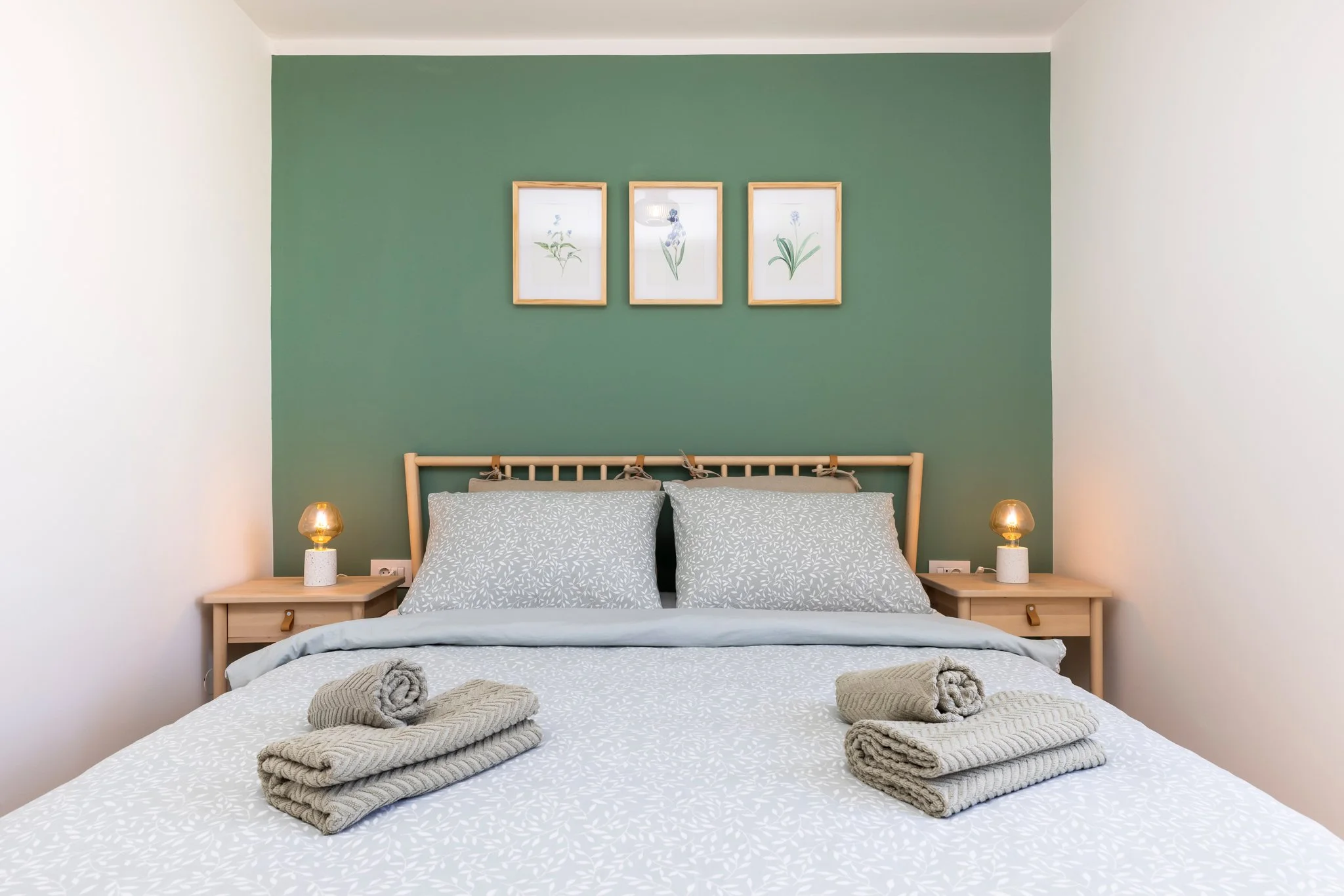 A neatly made bed with white and gray patterned bedding, two pillows, and rolled towels on top. Two bedside tables with small lamps are on either side of the bed. A green accent wall with three framed botanical prints behind the bed.