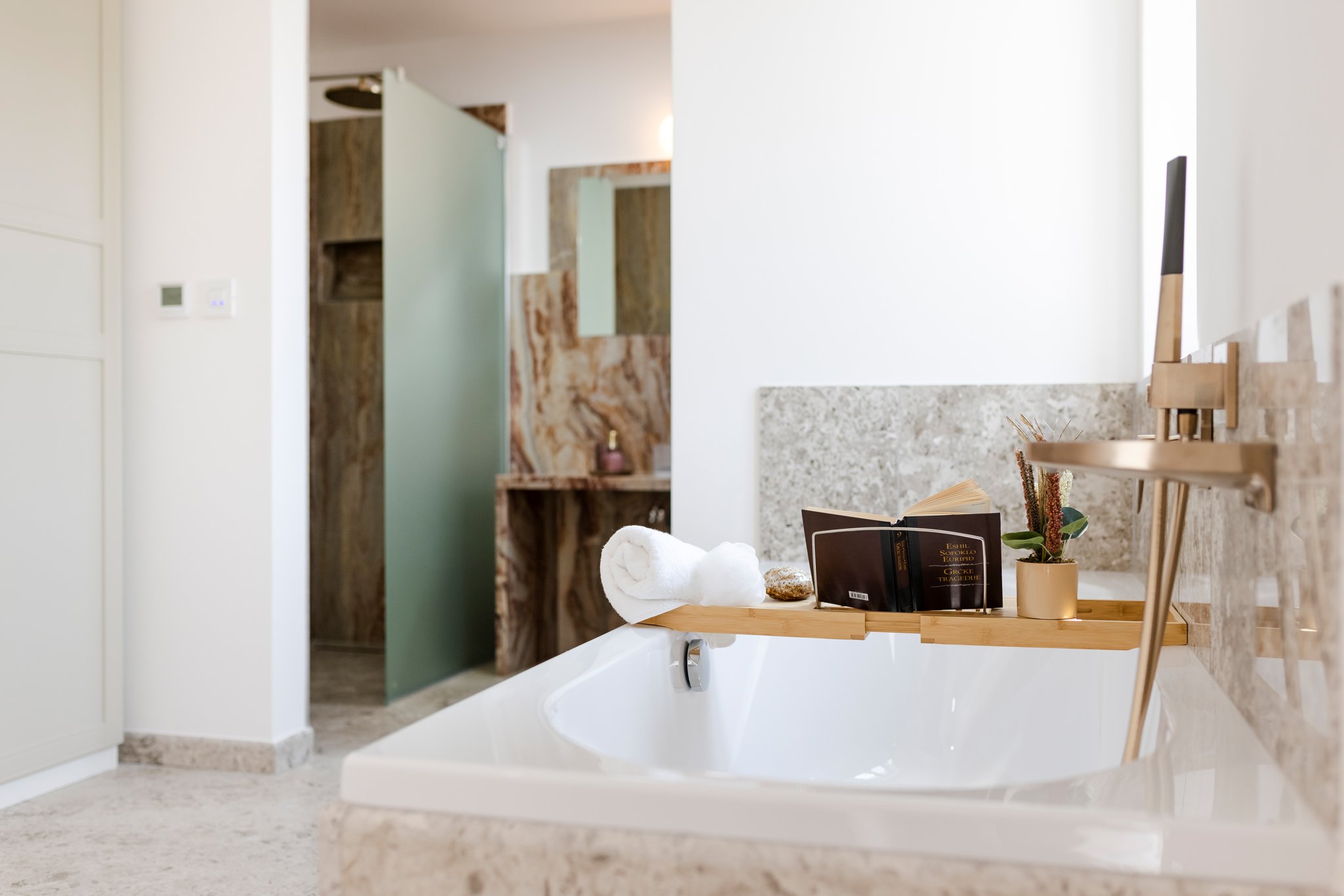 Bathroom with a white bathtub, wooden shelf with rolled towel, a book, a potted plant, and a soap dispenser, beige granite countertop, wall with a mirror, in a modern design.