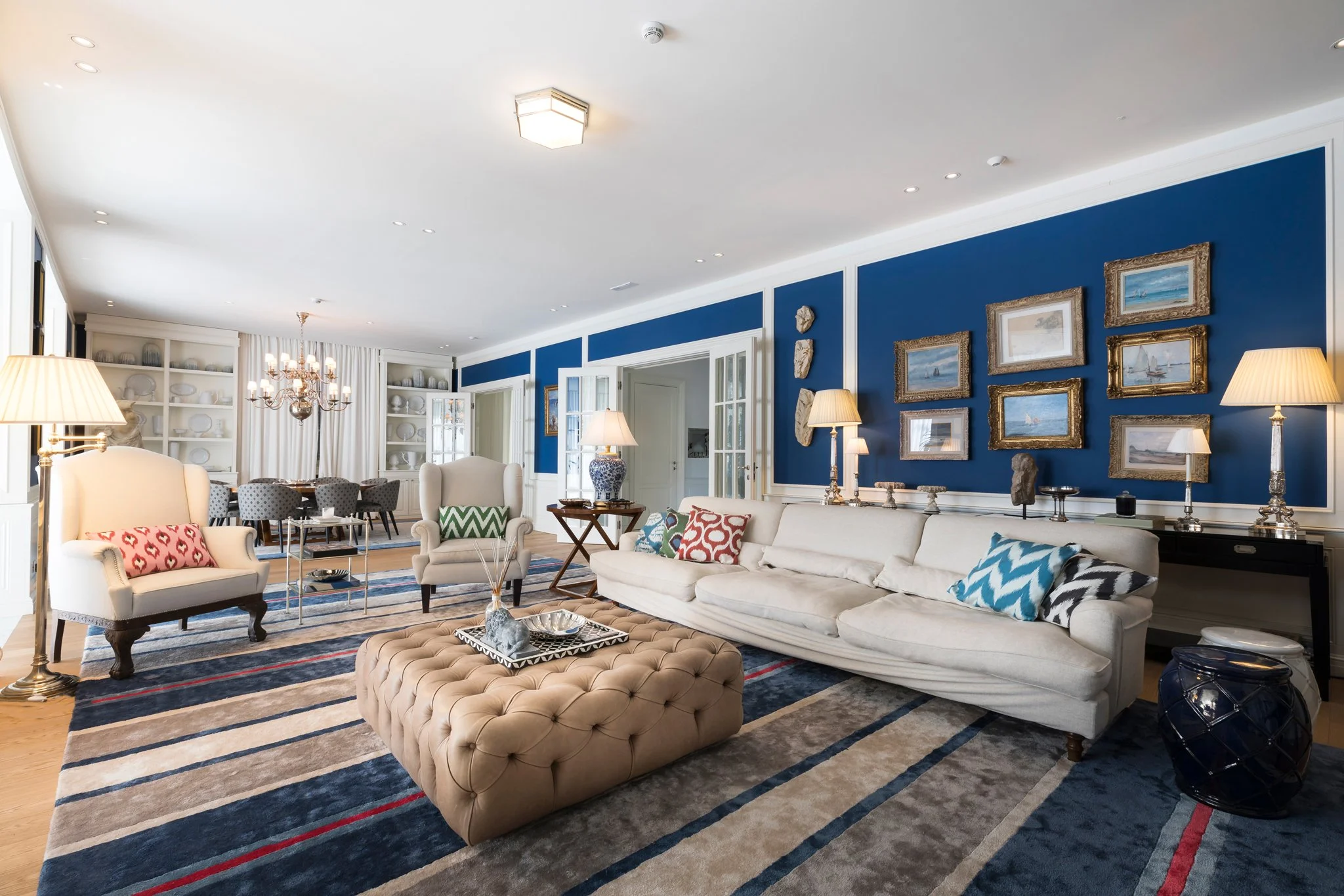 Living room with white sofas, patterned cushions, a tufted ottoman, a blue wall with framed paintings, a dining area with a chandelier, and lamps.