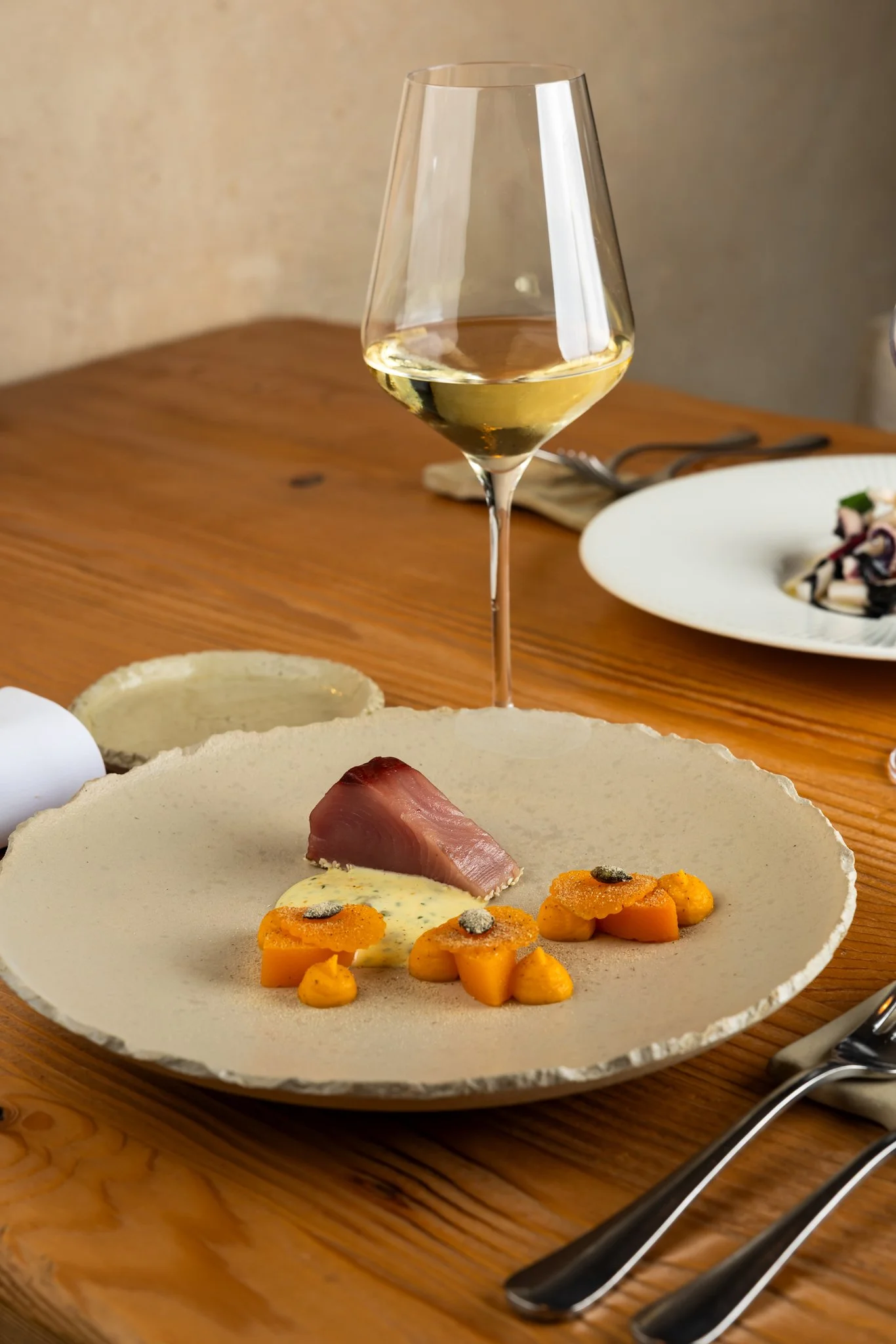 A plate of food with slices of fish, orange vegetables, and a yellow sauce, accompanied by a glass of white wine on a wooden table.