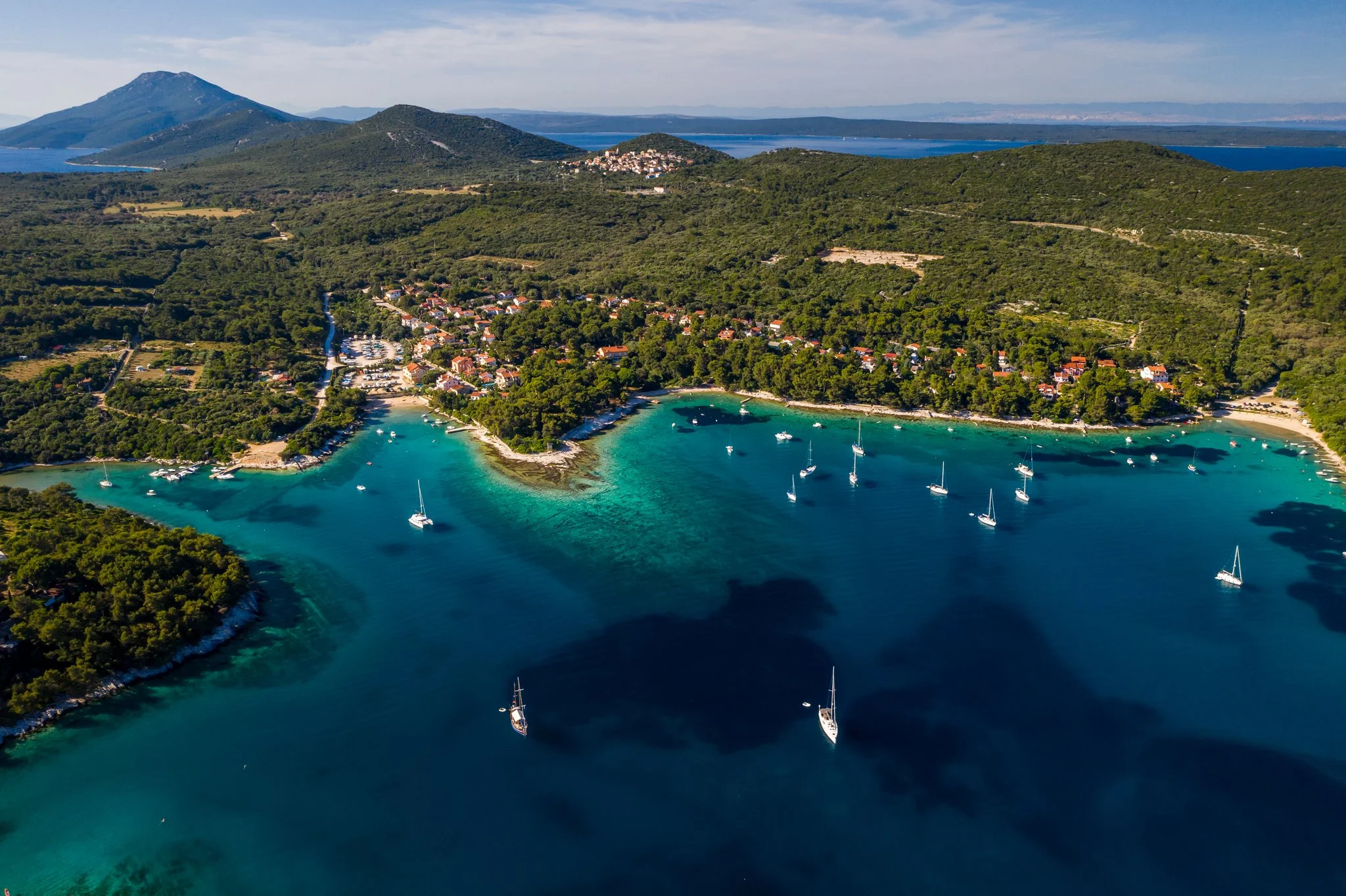 Aerial view of a coastal village with boats anchored in turquoise waters, surrounded by green hills and mountains.