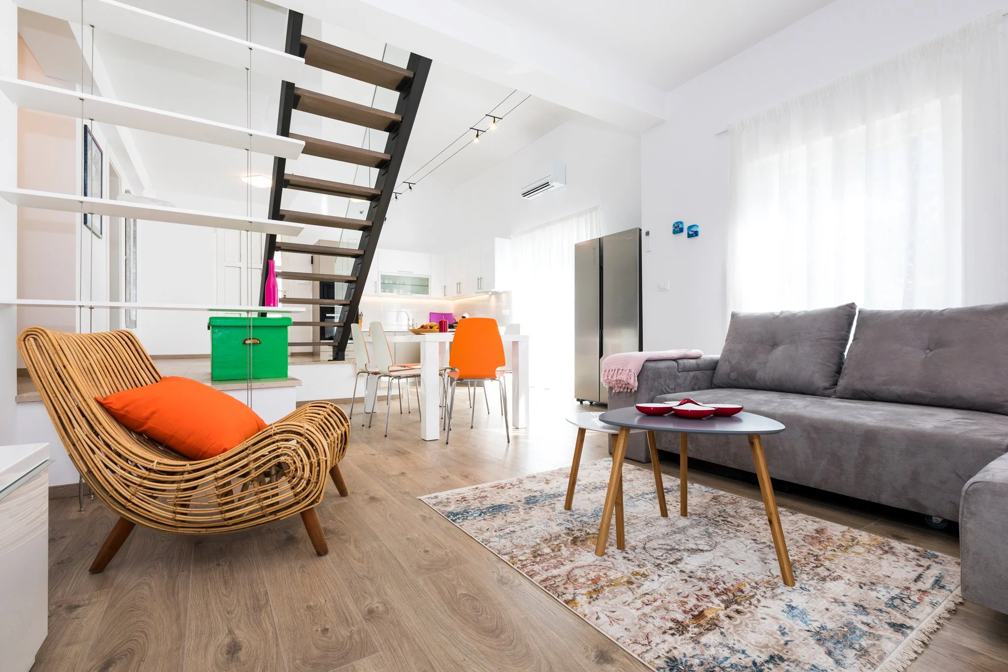 Interior of a modern, open-plan living room and dining area with white walls, light wooden flooring, a gray couch, a patterned rug, a small wooden coffee table with red bowls, and a rattan armchair with an orange cushion. Part of a staircase with woo
