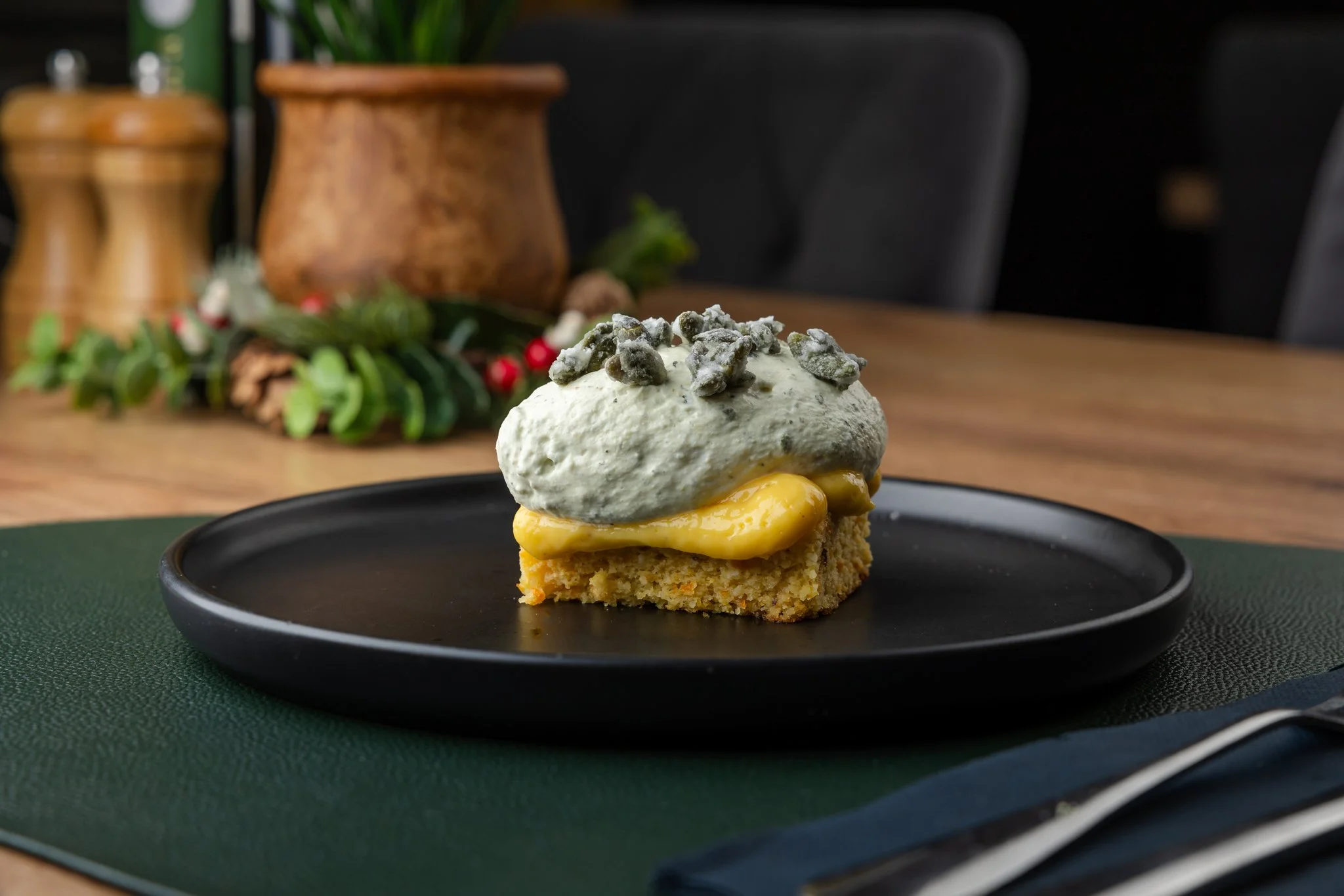 A dessert on a black plate with a yellow cake base topped with yellow custard, gray whipped cream, and garnished with gray edible seeds, set on a green placemat with a Christmas decoration in the background.