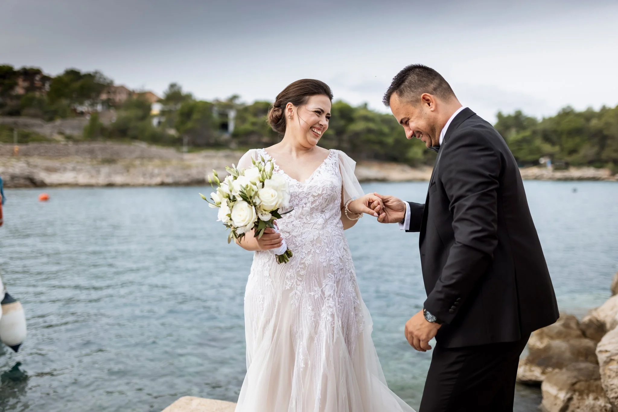 A bride and groom smiling and holding hands by the water, with a scenic rocky shoreline and trees in the background.