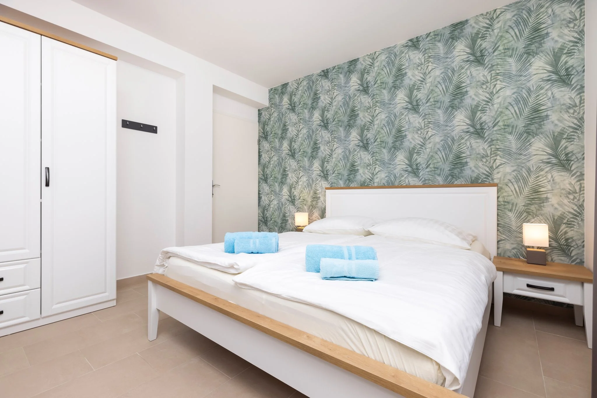 A modern bedroom with a white bed, tropical leaf wallpaper accent wall, two blue towels on the bed, side tables with lamps, and white closet doors.