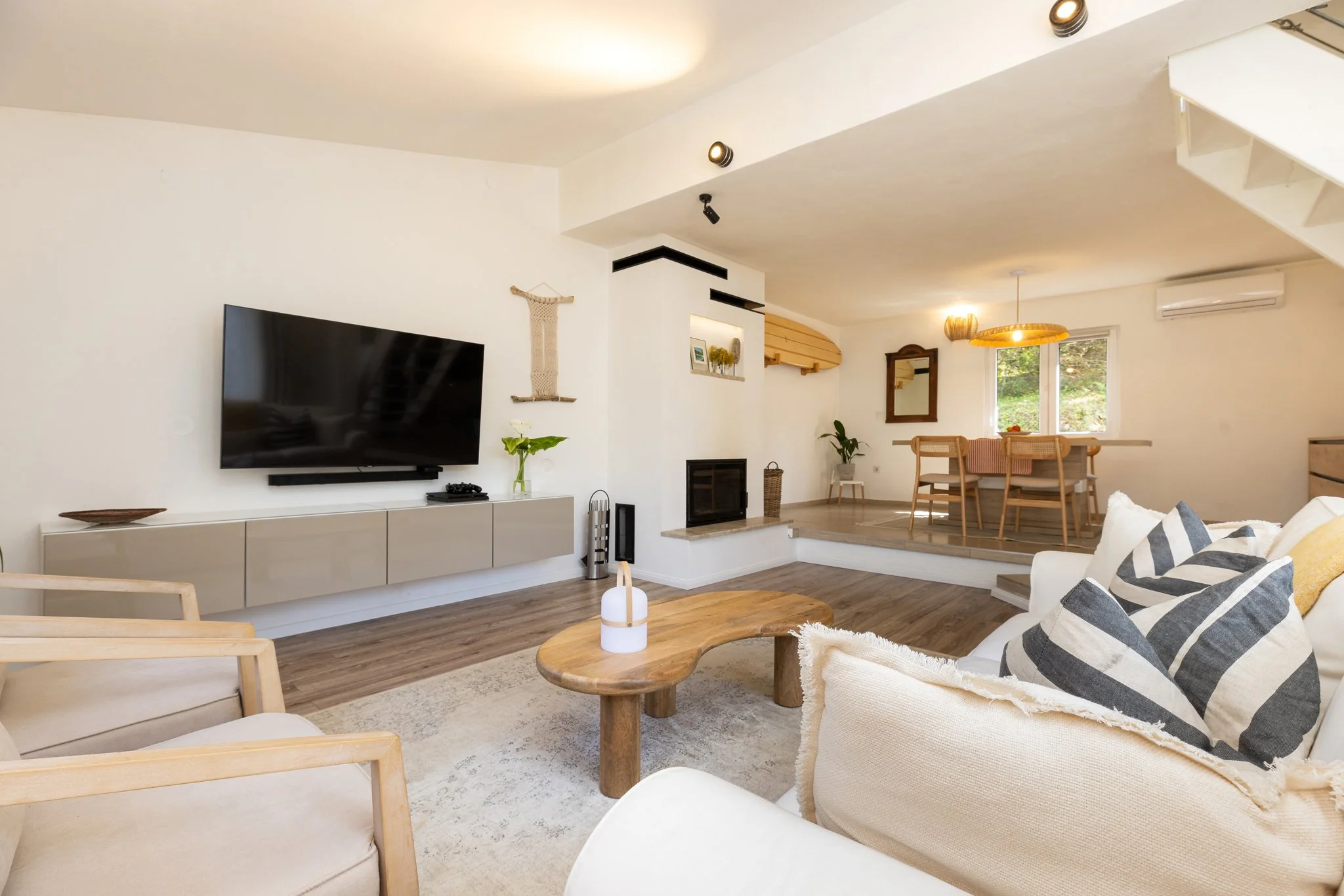 Open-concept living and dining area with white walls, wooden floors, a mounted flat-screen TV, beige couches with striped pillows, a wooden coffee table, potted plants, and a window showing trees outside.