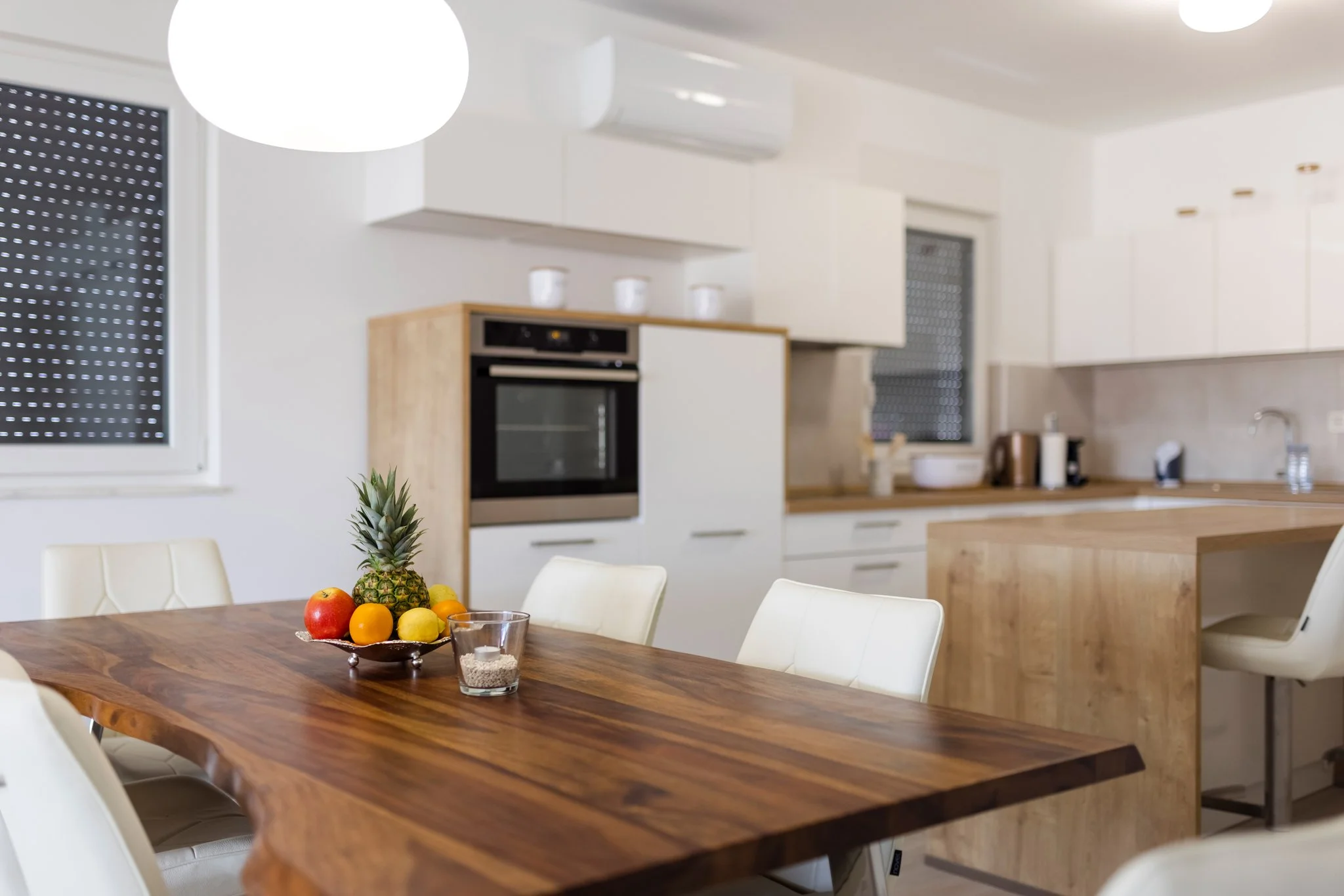 A modern, minimalist kitchen with white cabinets, a built-in oven, and a wooden countertop. There is a dining table with a fruit bowl containing pineapple, apples, oranges, and lemons, along with a glass of water and a candle. White chairs surround t