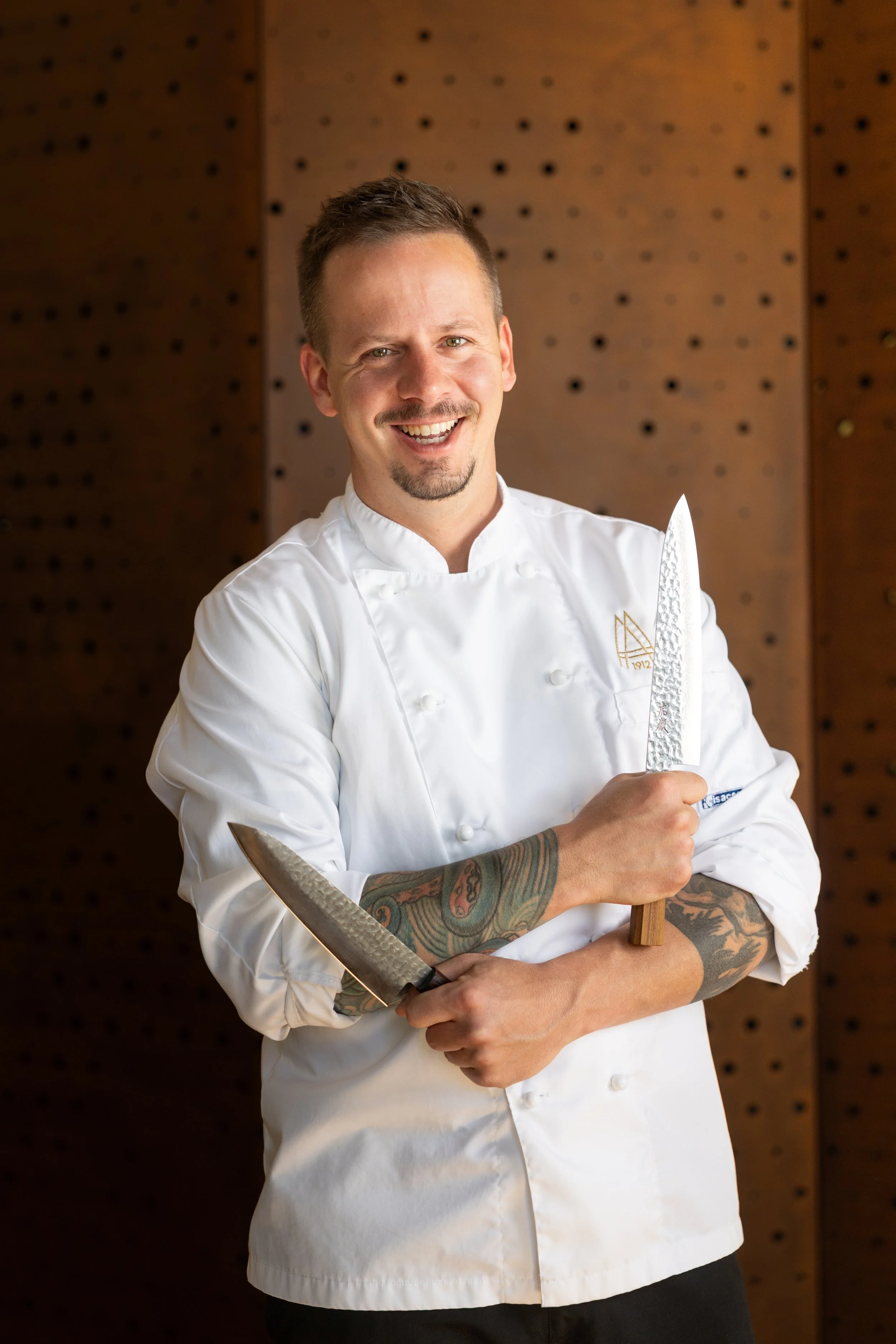 A smiling male chef with tattoos on his arm, wearing a white chef's coat, holding a large knife with both hands, standing in front of a wooden wall with small holes.
