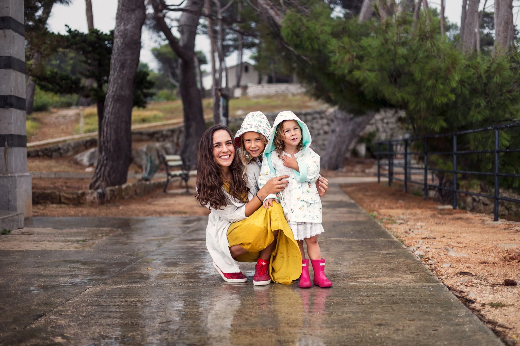 A woman with long brown hair smiling while kneeling, holding two young girls on a wet outdoor path with trees and a fence in the background. The girls are wearing raincoats and rain boots, one in pink and the other in patterned rain gear. The scene a