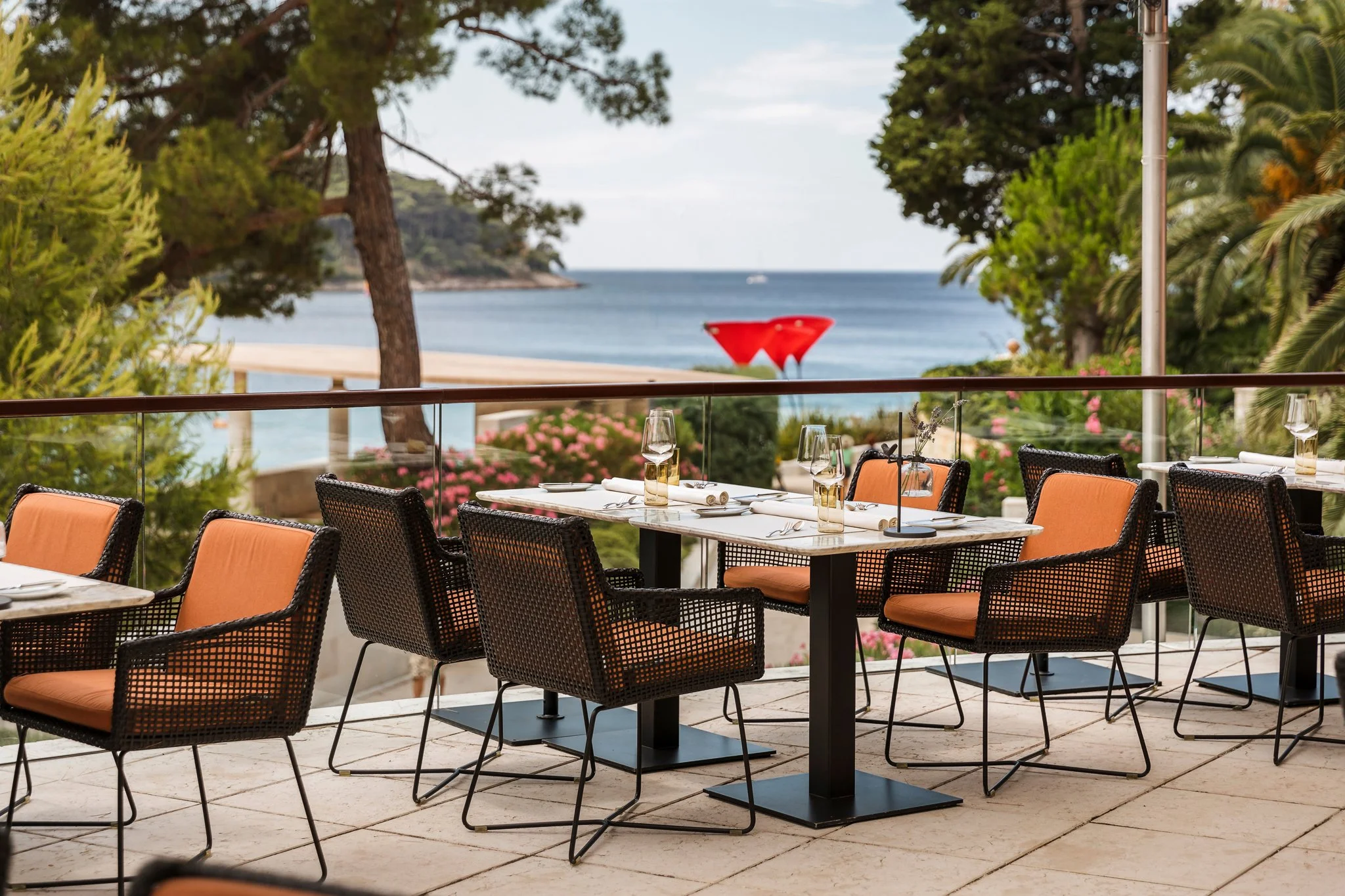 Outdoor restaurant patio with tables set for dining overlooking a beach, framed by trees and greenery. Alfred Keller in Mali Lošinj, Boutique Hotel Alhambra.
