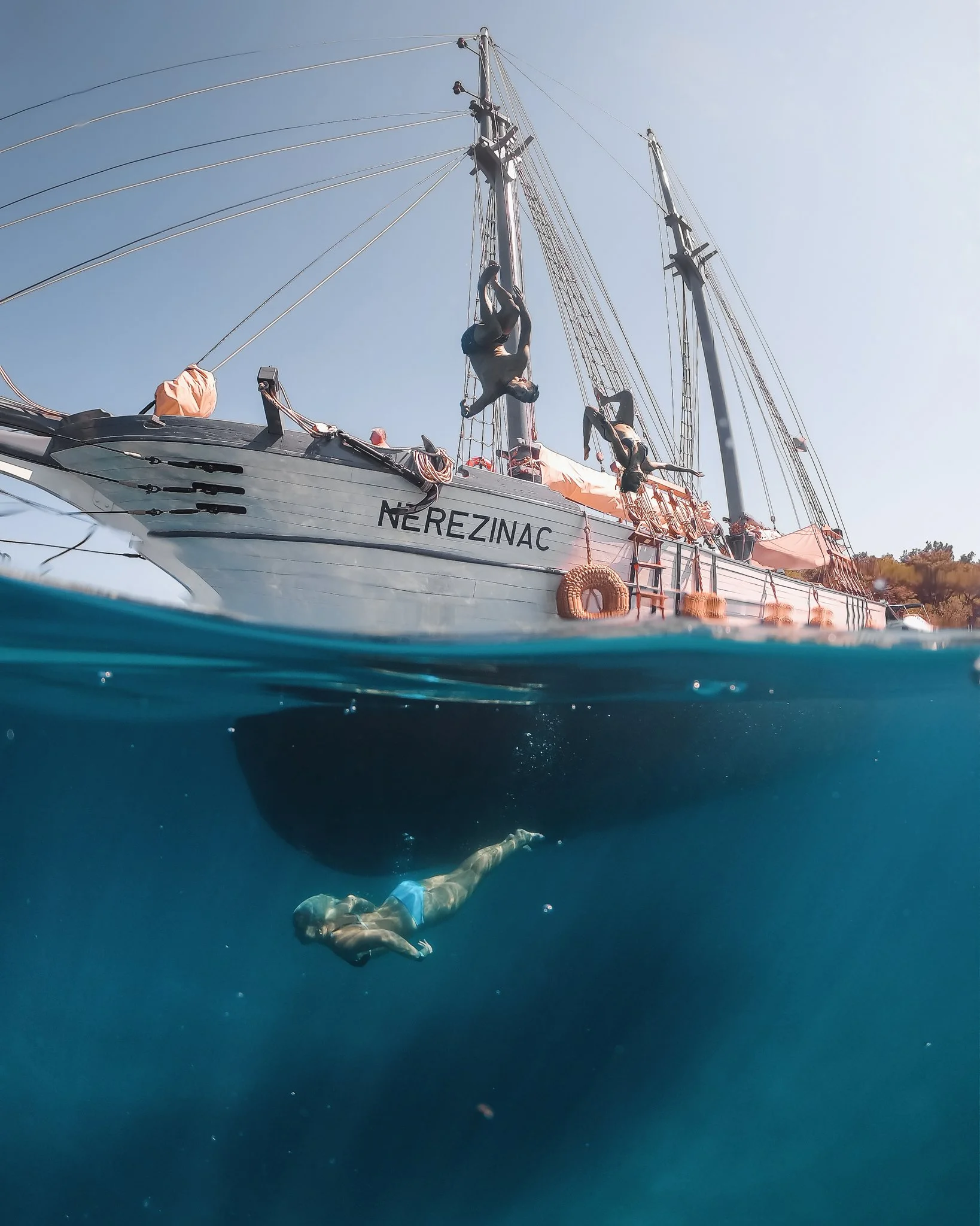 A person swimming underwater near a large sailboat named 'NEREZINAC' with people on deck, and the ocean visible both above and below the water.