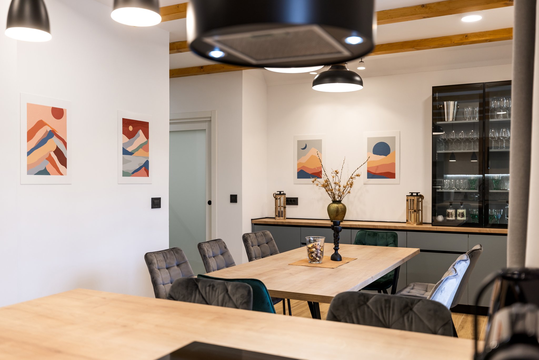 A dining room with a wooden table surrounded by gray upholstered chairs, a vase with dried flowers, and a black candle holder, with framed abstract mountain landscape art on the white walls and black pendant lights hanging from a wooden-beamed ceilin