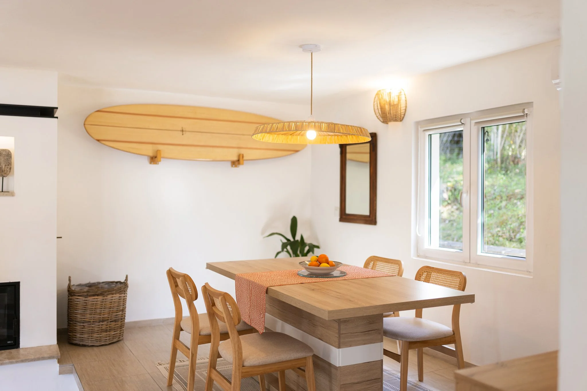 A dining area with a wooden table, six chairs, a bowl of fruit, a large window, and a light fixture. A surfboard is mounted on the wall.
