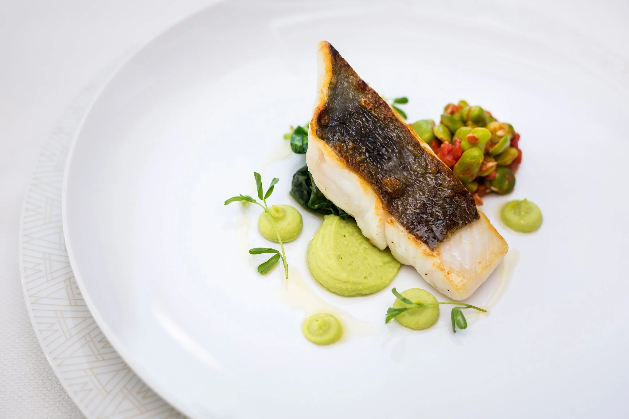 A plated dish featuring a piece of white fish with crispy skin, accompanied by green pea puree, a small green vegetable, and a colorful spicy tomato relish, garnished with microgreens.