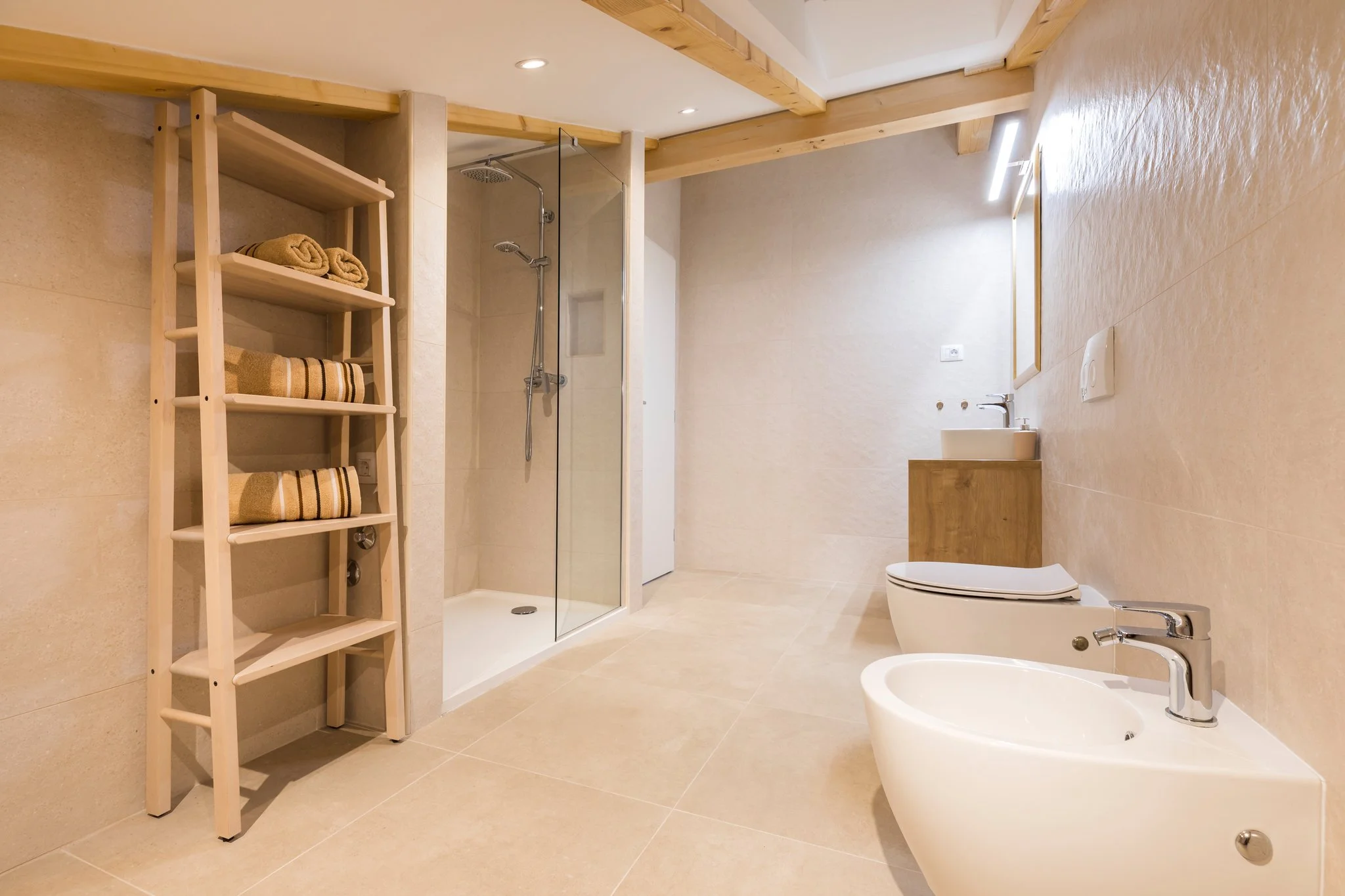 A modern bathroom with beige tiled walls and floors, a glass-enclosed shower, a wooden shelf with towels, a bidet, and a toilet, all with minimalist fixtures and wooden accents.
