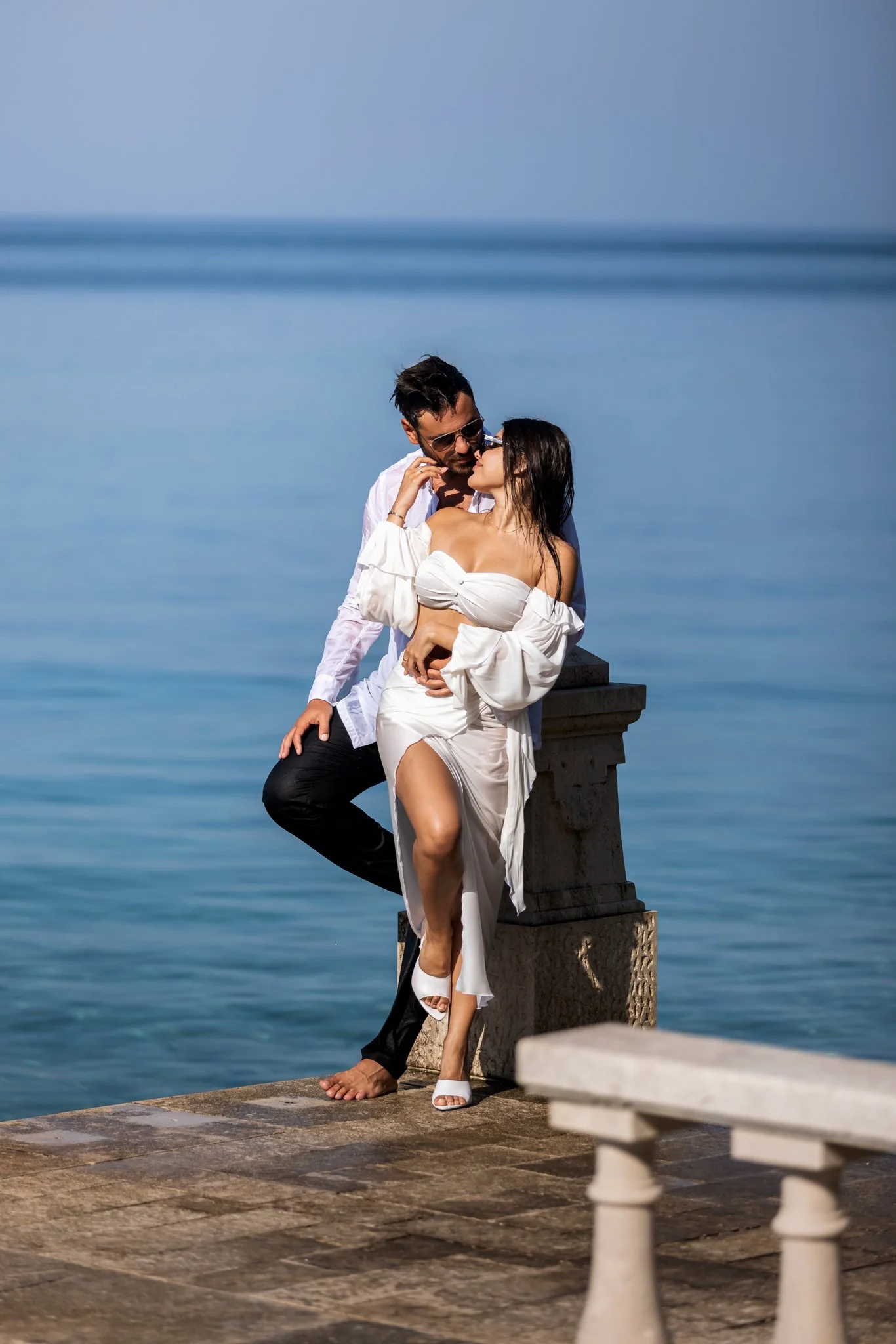 A man and woman standing close together on a waterfront promenade, with the ocean and a pier in the background. The woman is dressed in white and high heels, and the man is in dark pants and a white shirt. They are sharing an intimate moment.