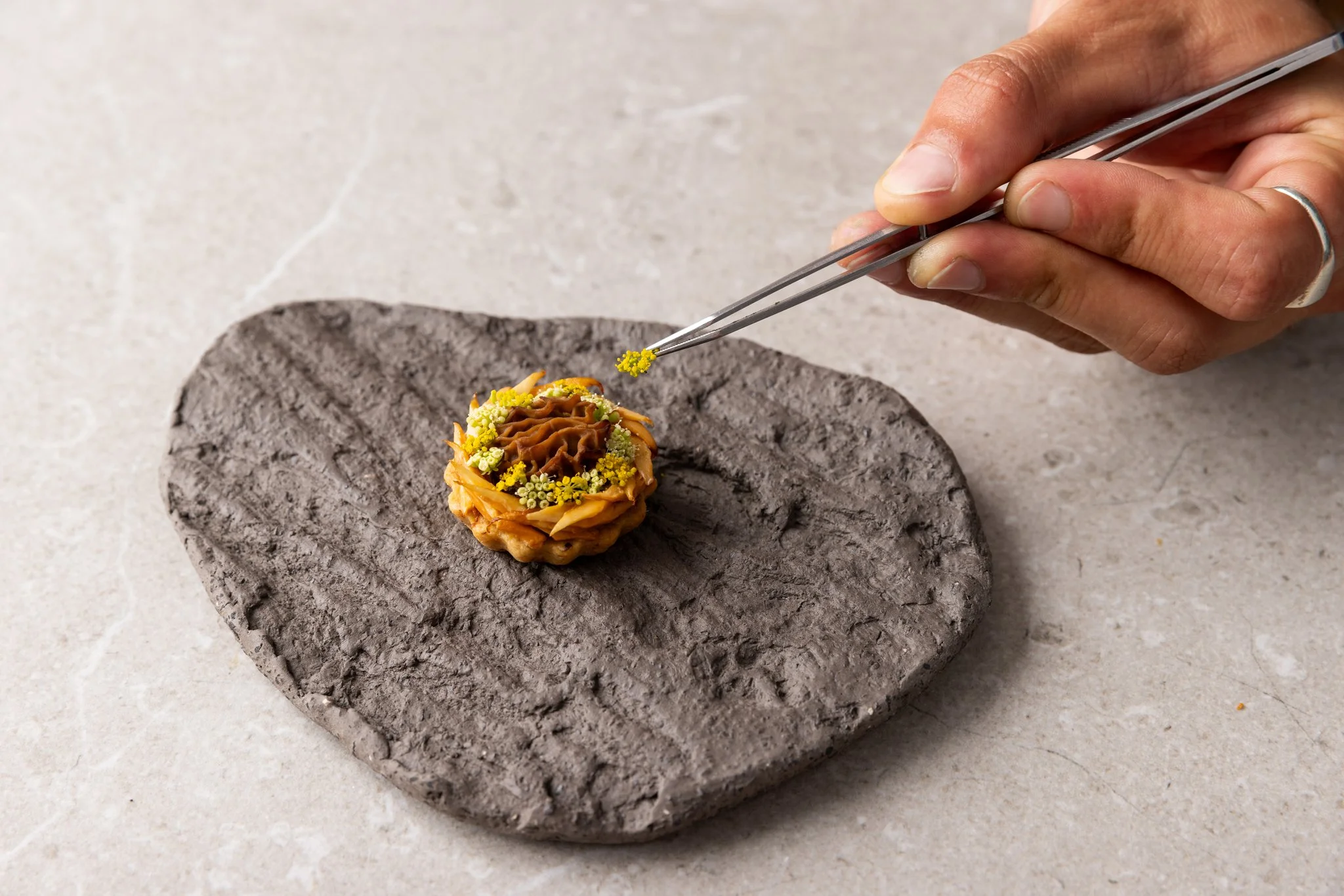 A person using tweezers to place yellow flower-like decorations onto a small, round, dessert that is decorated with various colorful toppings, on a dark stone slab.