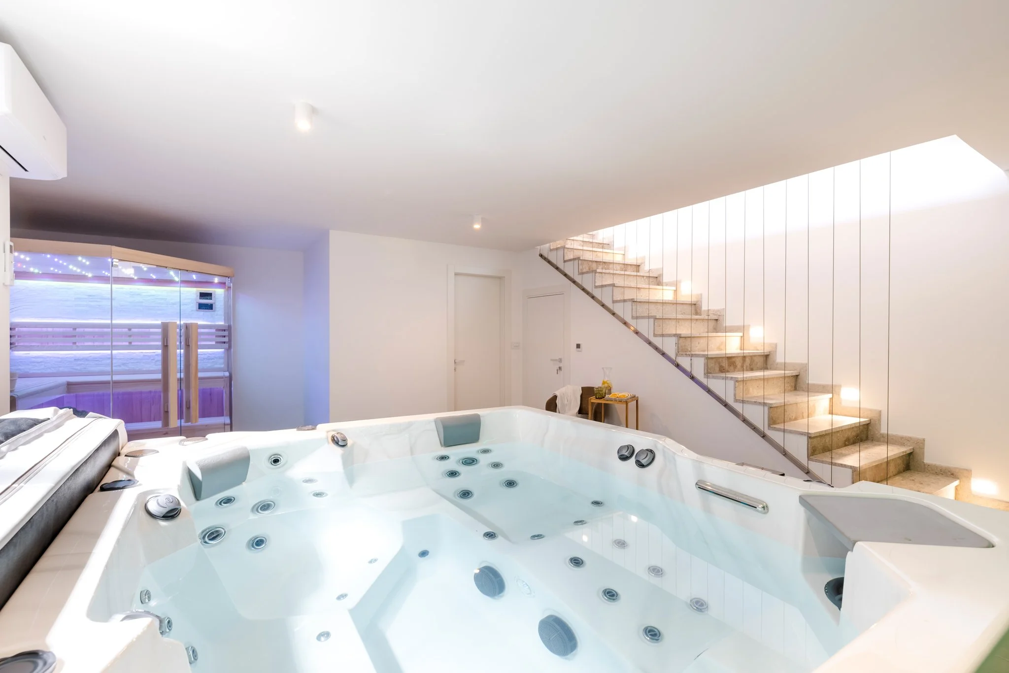 A modern indoor space with a hot tub, staircase, and large window with blinds and string lights.