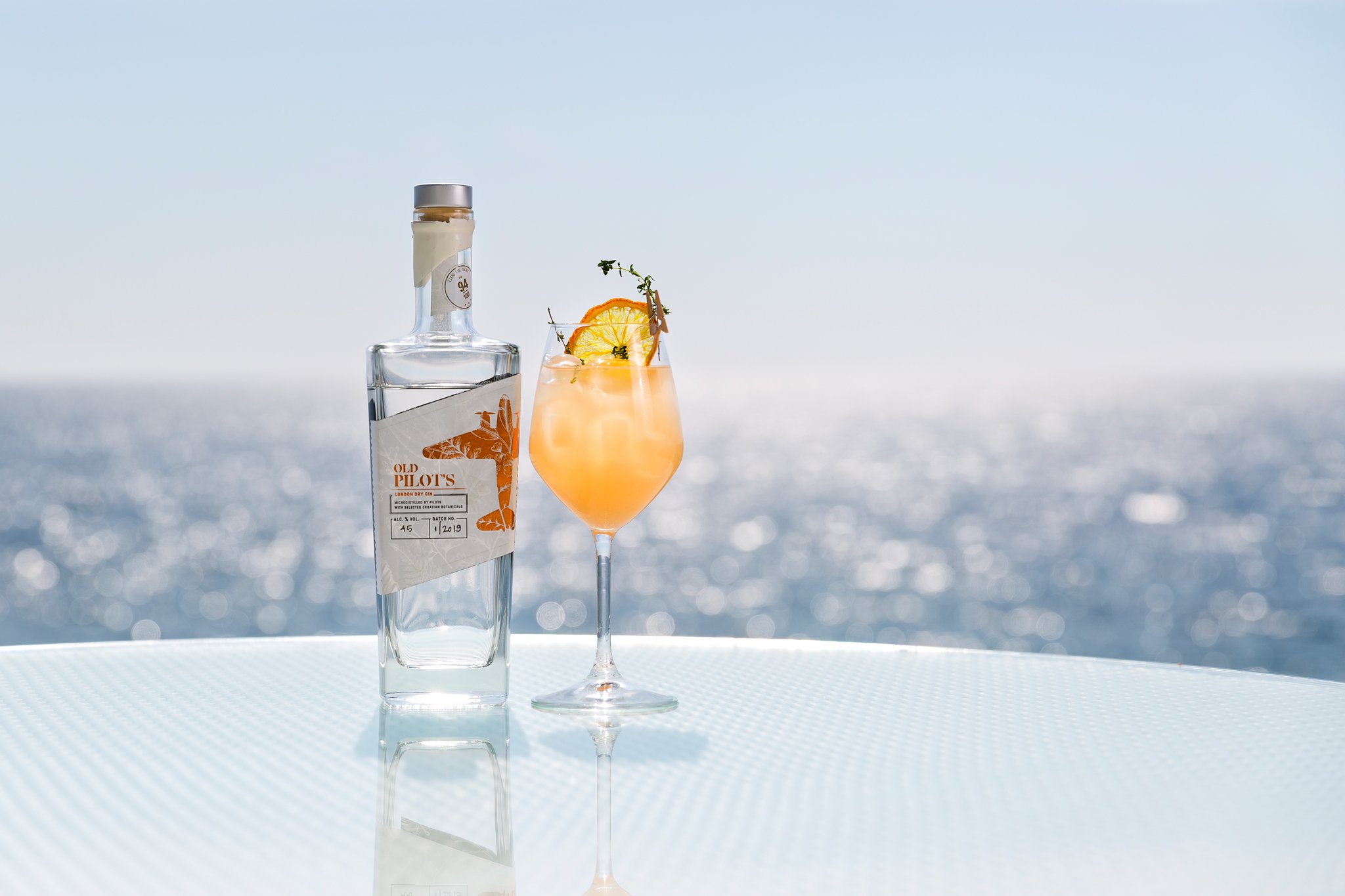 A clear bottle of Old Pilot's gin and a cocktail glass with a citrus garnish and herbs, set against a blurred seaside background with sunlight reflecting on water.
