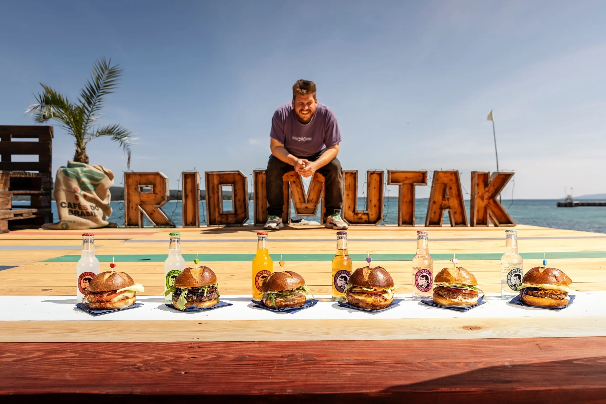 A man smiling and squatting behind a sign that spells 'ROCKTALK' near a body of water. In the foreground, five gourmet burgers with different toppings and sauces are placed on a white table, each with a small branded flag, alongside bottles of bevera