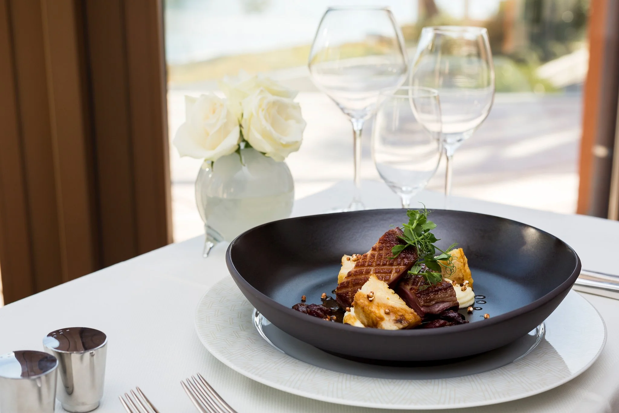 A black bowl with a dessert containing layers of ice cream, chocolate, and garnished with greens, set on a white plate. In the background, a small white vase with white roses, empty wine glasses, and a window with a blurred outdoor view are visible.