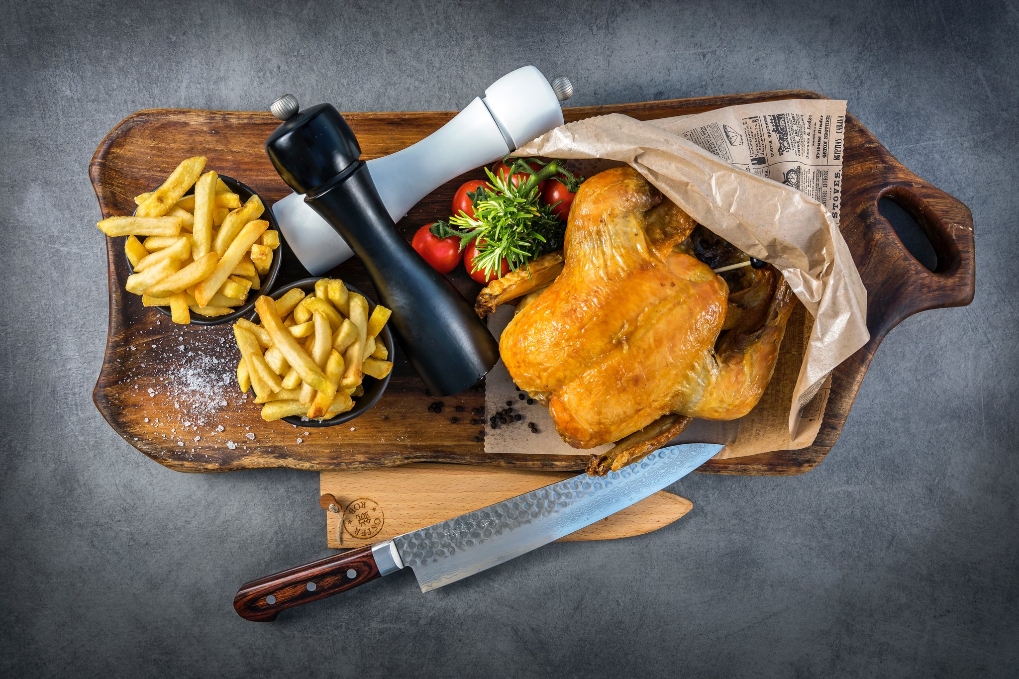 Roasted whole chicken on a wooden cutting board, with a side of French fries in a small black bowl, cherry tomatoes, and a sprig of green herb, on a gray countertop, with a chef's knife and a meat hammer.