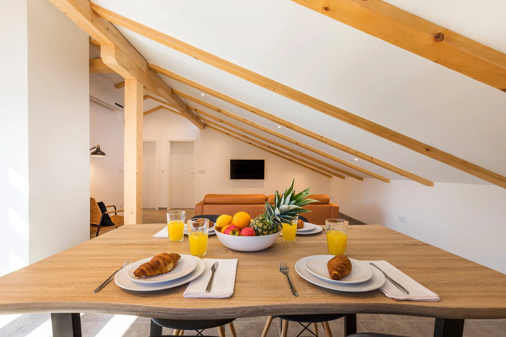 A dining table set with plates, croissants, glasses of orange juice, and a bowl of fruit, in a room with sloped ceiling beams and a TV on the wall.