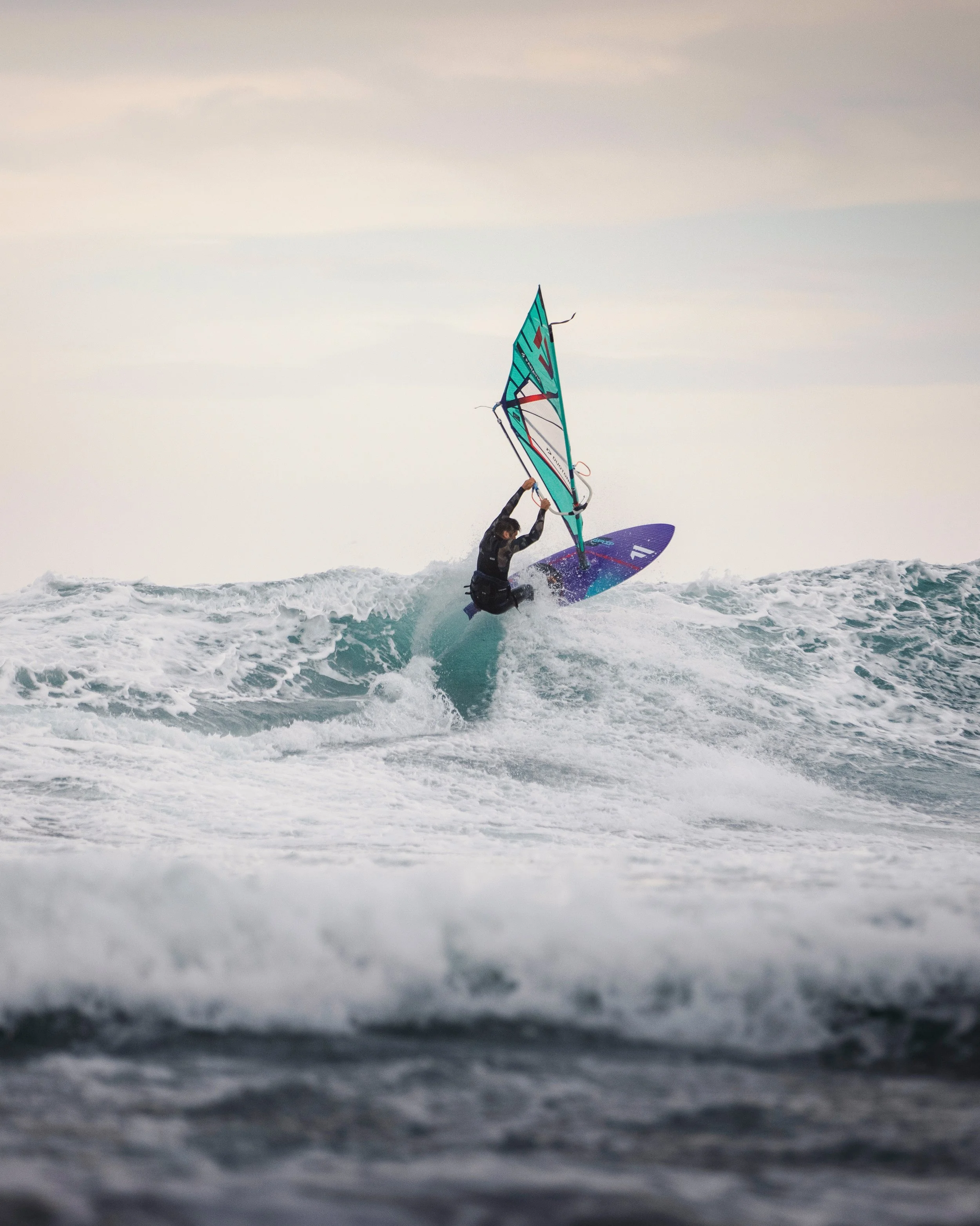 A person windsurfing on the ocean during cloudy weather, with a colorful sail and purple board.