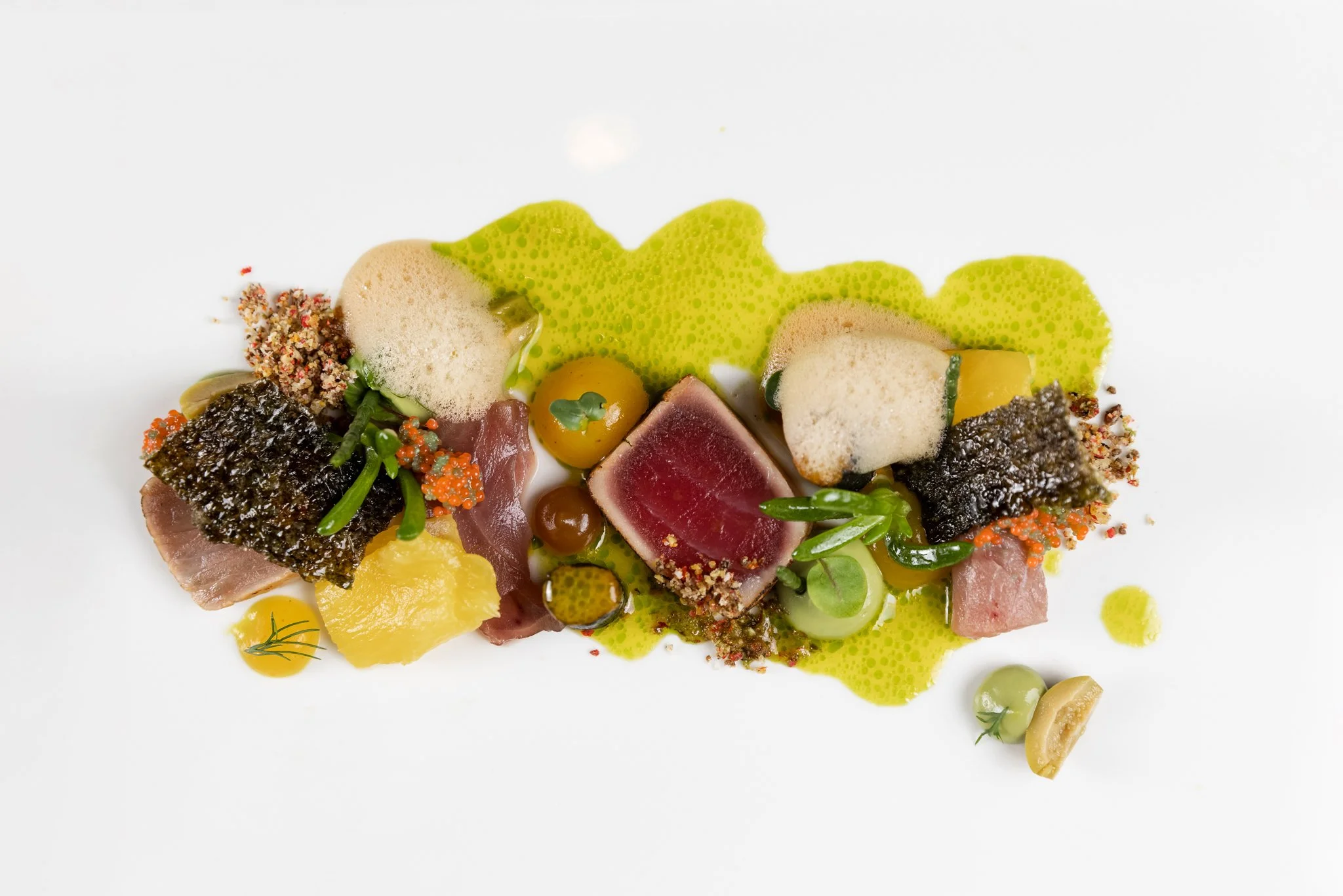 Chef's plated gourmet dish with seared tuna, green foam, pickled vegetables, and microgreens, garnished with foam and sauce on a white background.