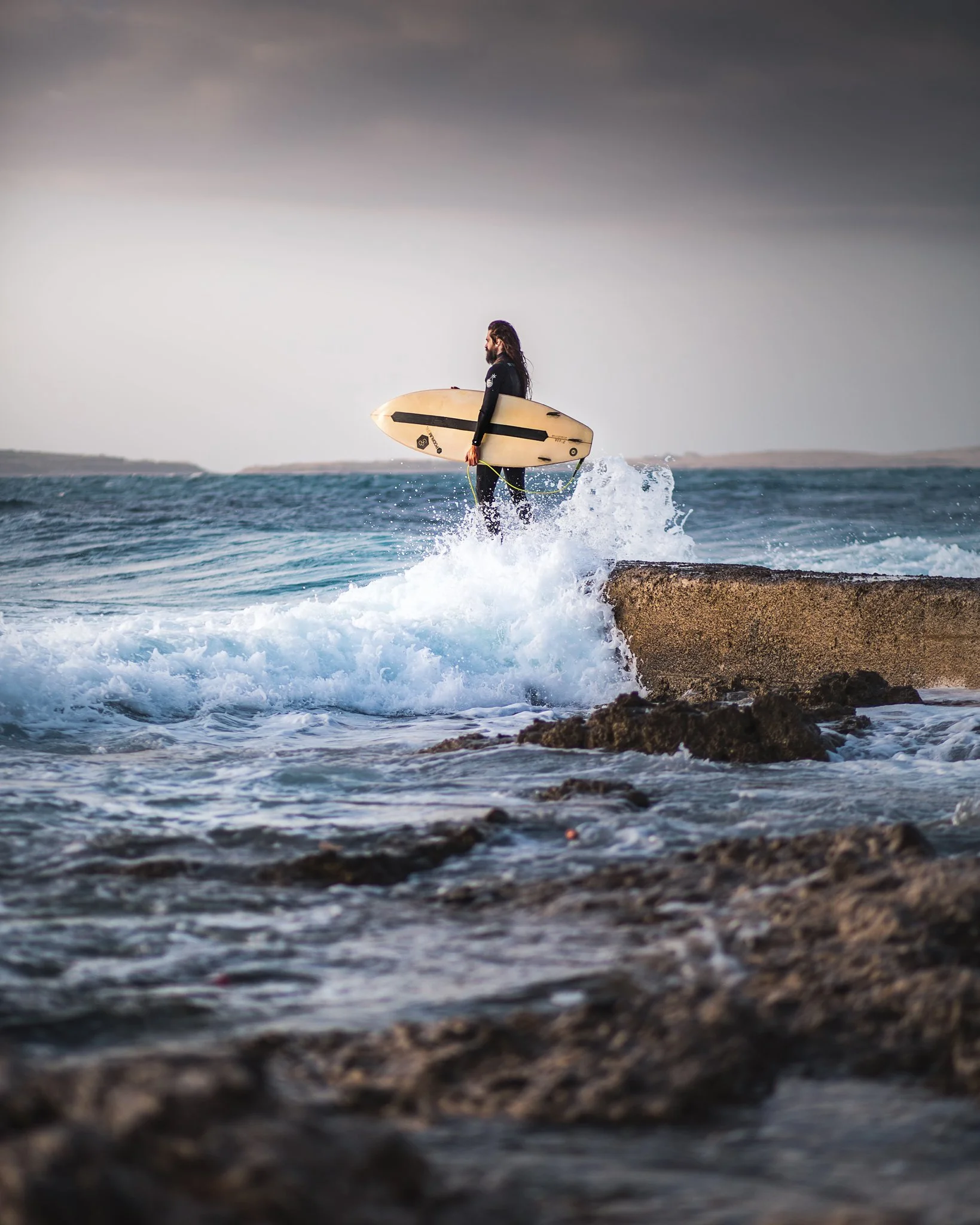 A man with long hair and a beard, dressed in a black wetsuit, carrying a surfboard and standing on rocks by the ocean, with waves crashing below and a cloudy sky overhead.