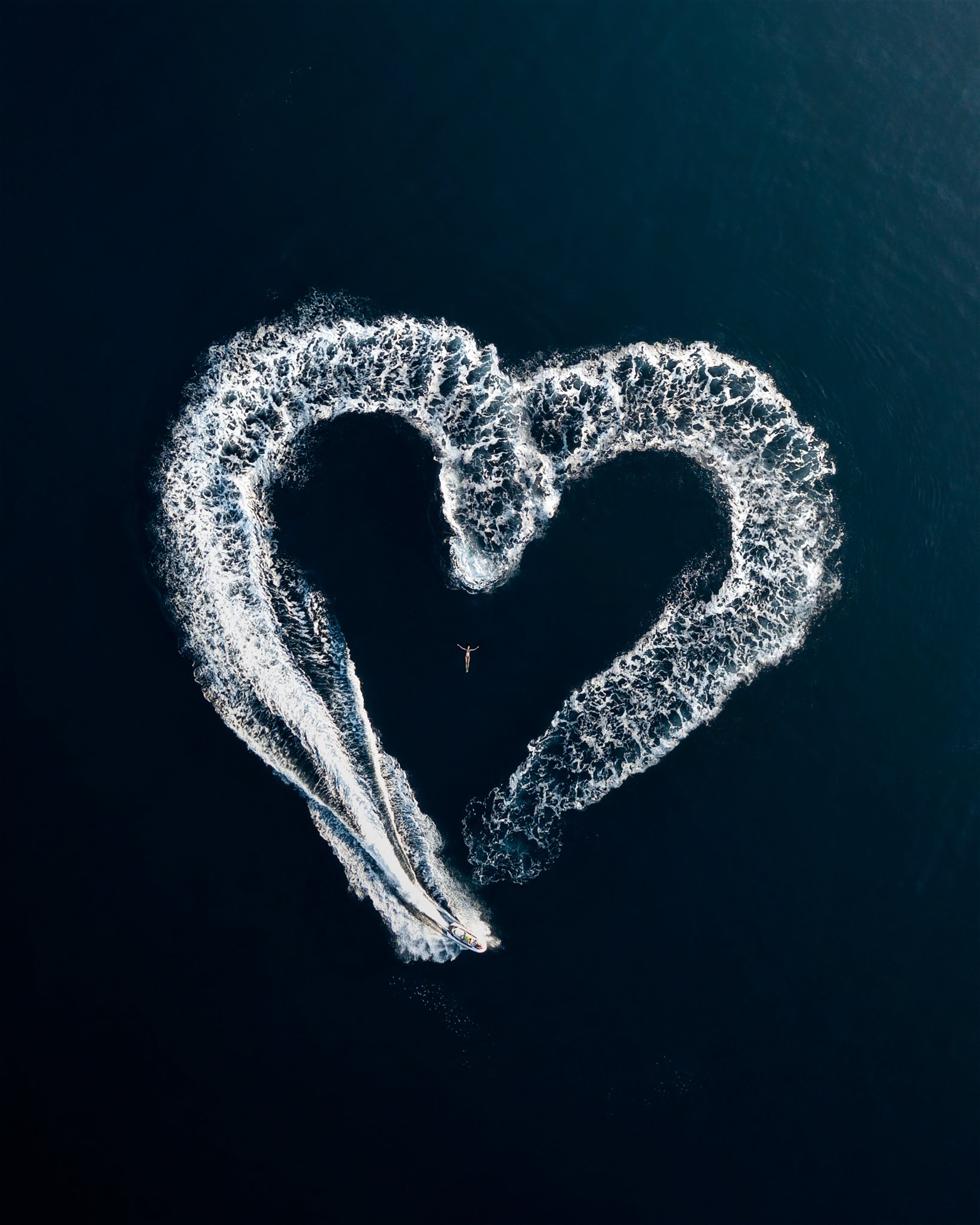 An aerial view of a person wakeboarding creating a heart shape in the water, surrounded by a trail of white foam.