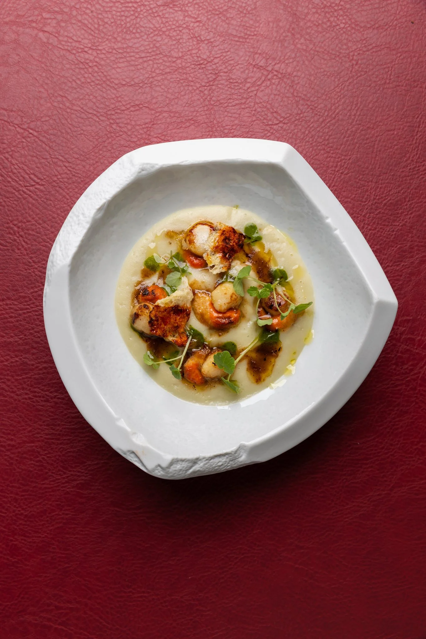 A white plate with grilled scallops and vegetables on a bed of creamy sauce, garnished with microgreens, on a red textured surface.