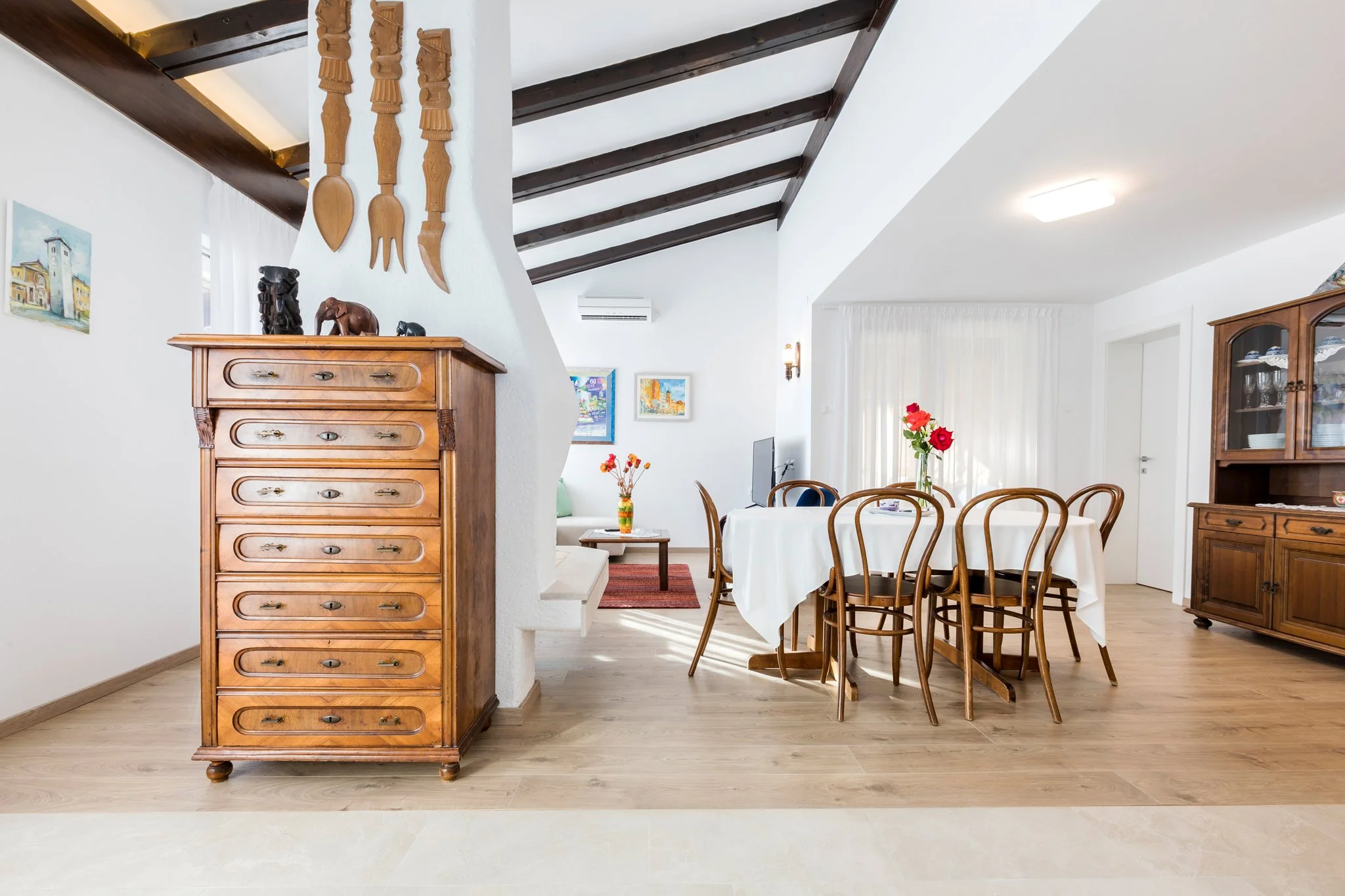 A bright, airy dining room with white walls and dark wooden ceiling beams. A round table with a white tablecloth and wooden chairs is centrally positioned, with a glass vase of red and orange flowers on it. An antique wooden cabinet with glass doors 
