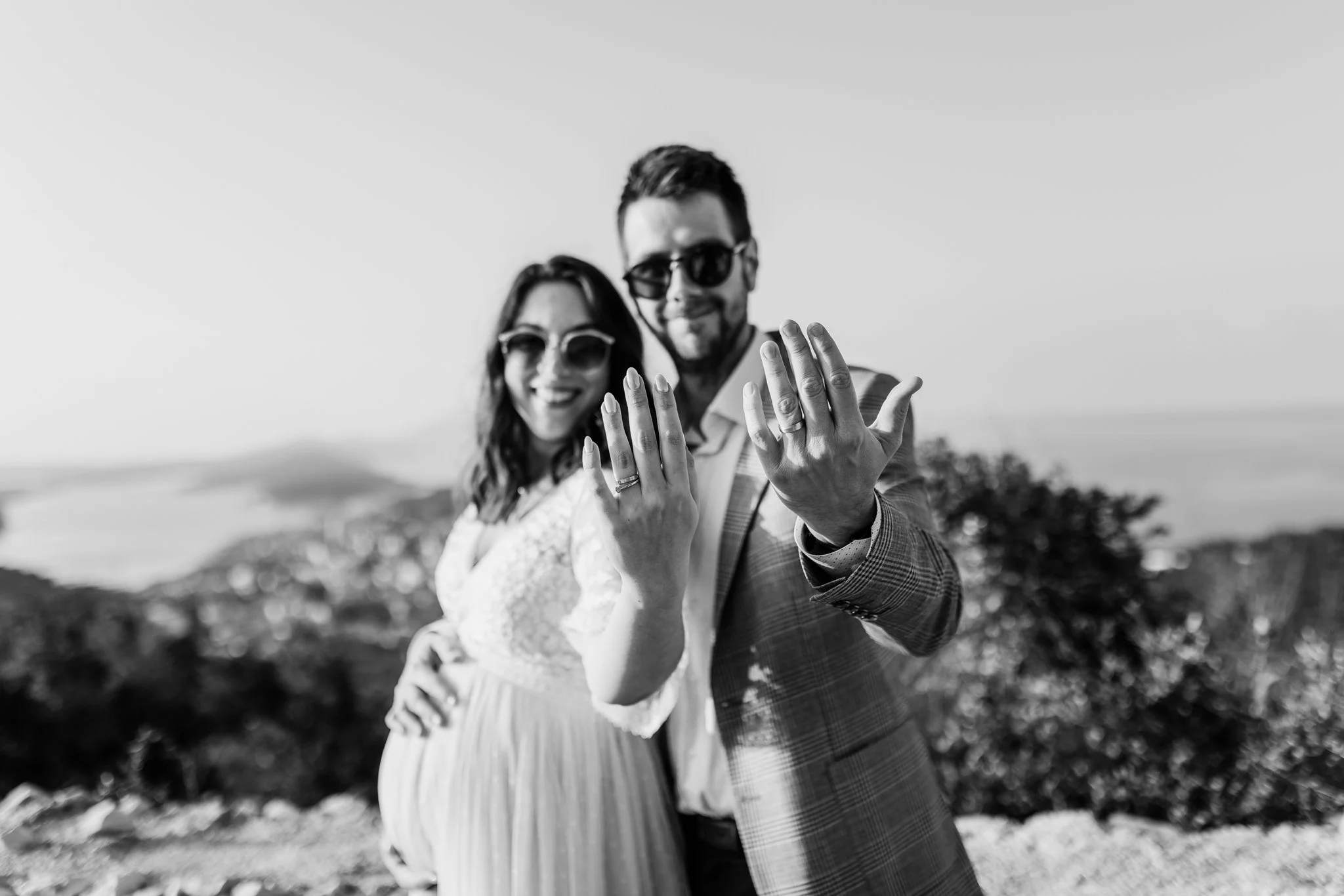 A smiling couple with sunglasses showing off their wedding rings, outdoors with a blurred natural background.