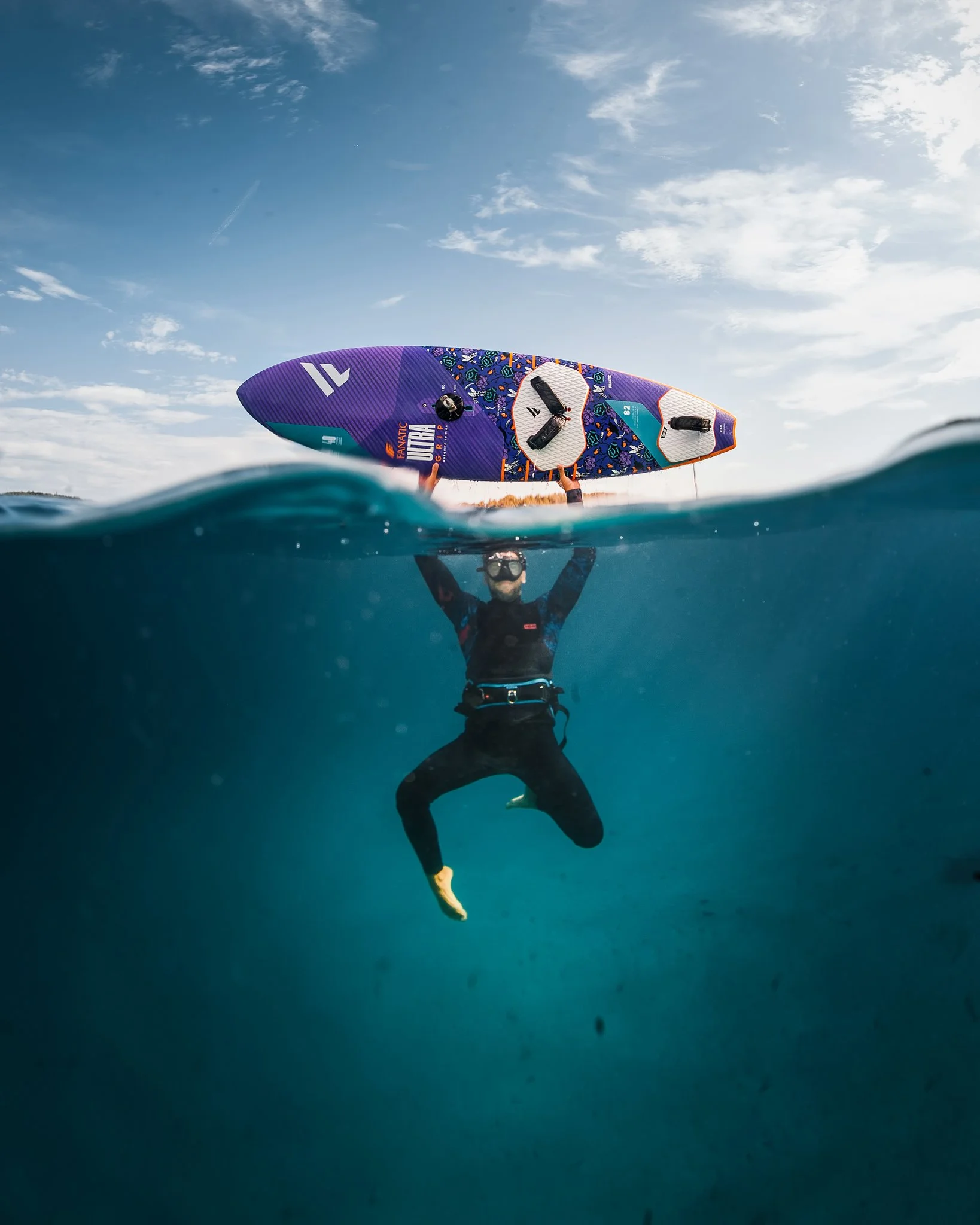 A person wearing a wetsuit standing in water, holding a colorful windsurfing sail above their head, with a clear sky and clouds in the background.