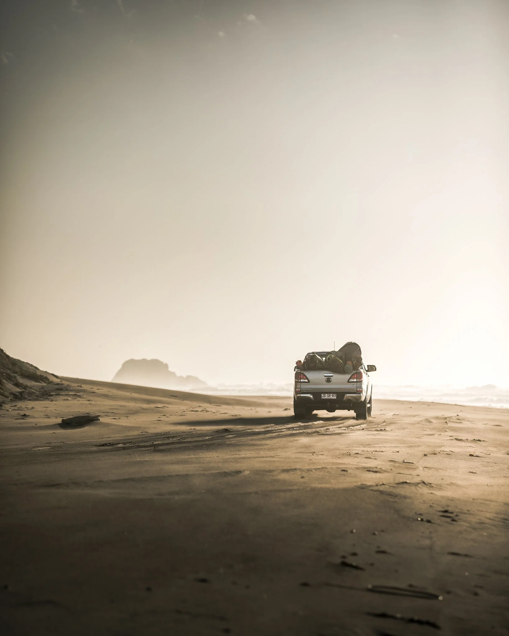 A car parked on a sandy beach with a large ocean wave crashing in the background during what appears to be early morning or late evening.